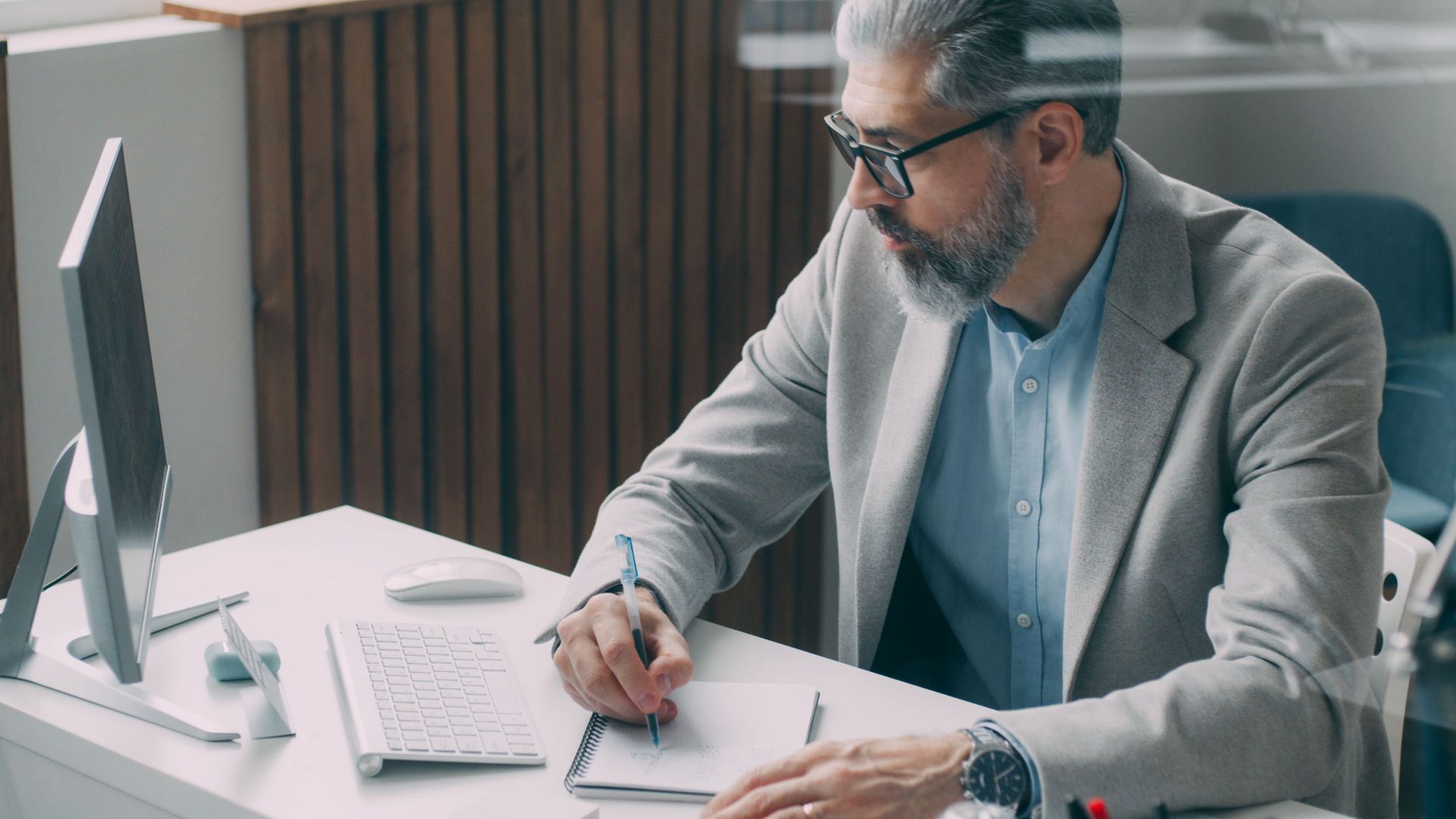 A mature businessman taking notes at his desk with a computer, wearing glasses and a suit jacket.