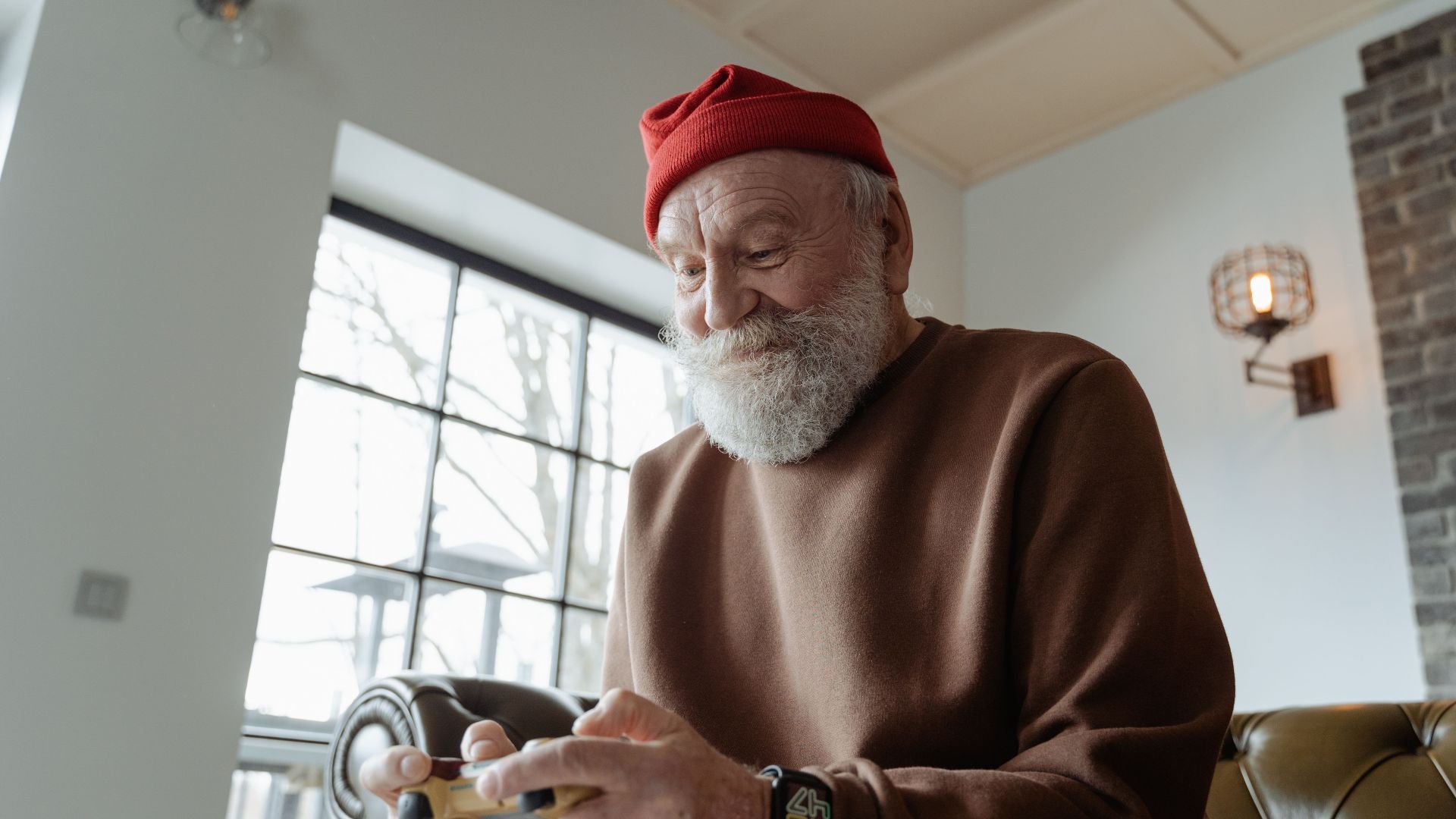Senior man with beard and red hat playing a video game indoors, enjoying leisure time.
