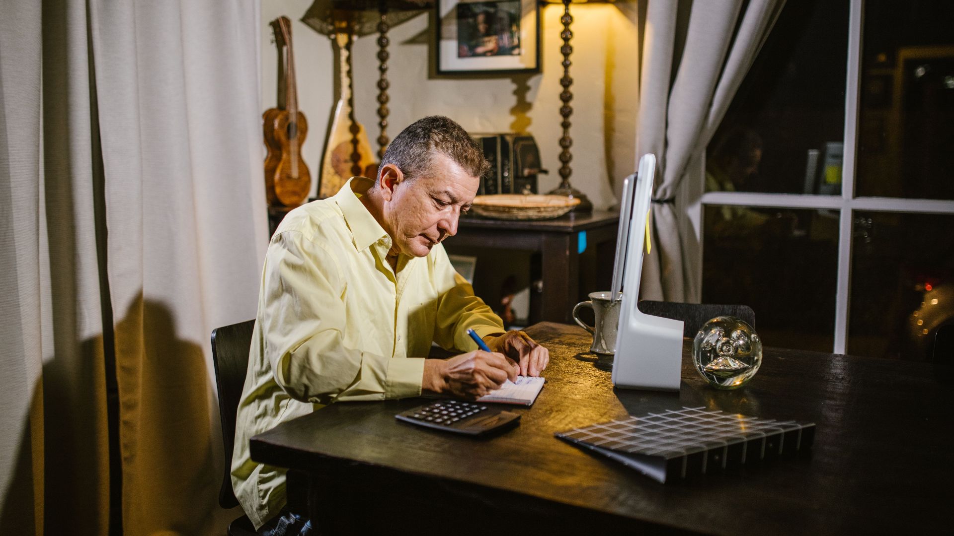 Senior man wearing yellow shirt writing notes at desk with lamp, calculator, and glass sphere nearby.