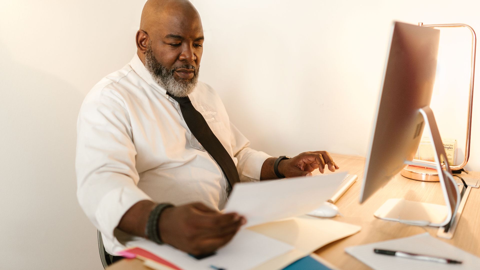 Bald man with beard in white shirt reviewing documents at desk with computer.