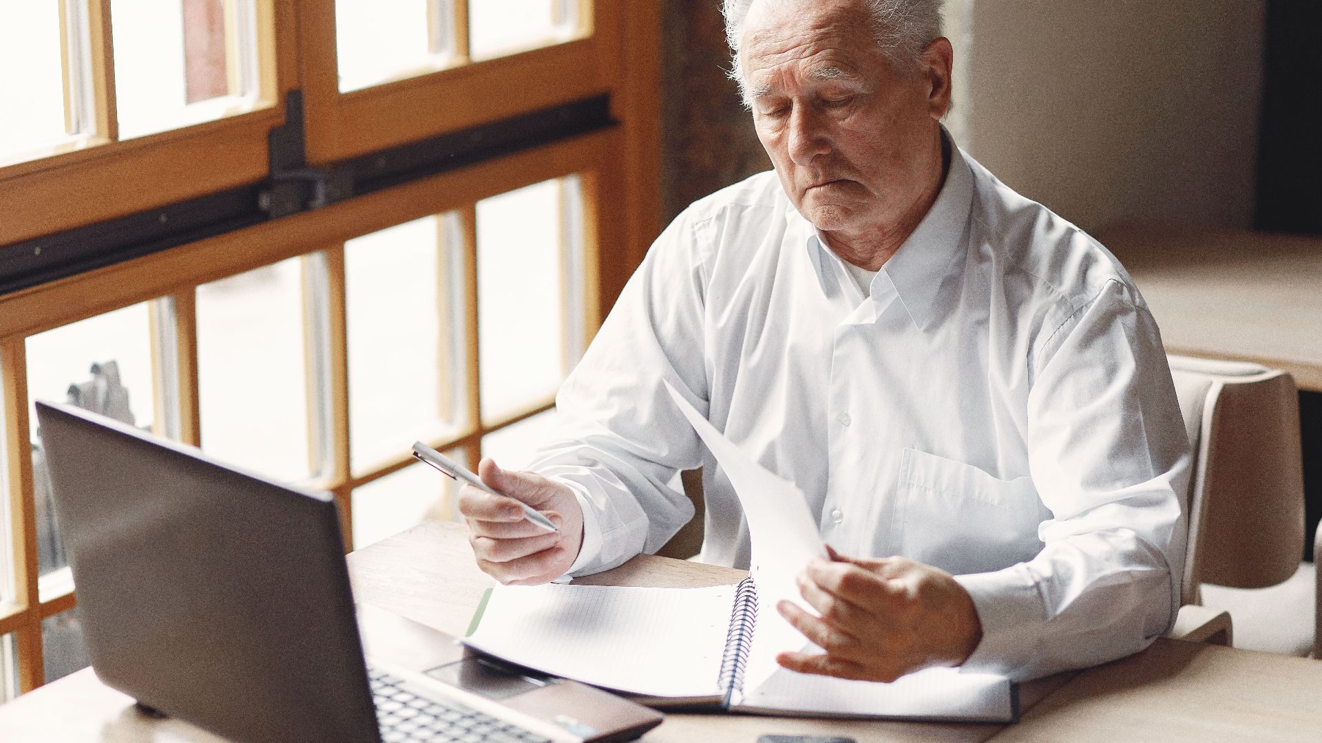 Serious gray haired aged man in white shirt sitting at table and browsing laptop while checking notes in modern office