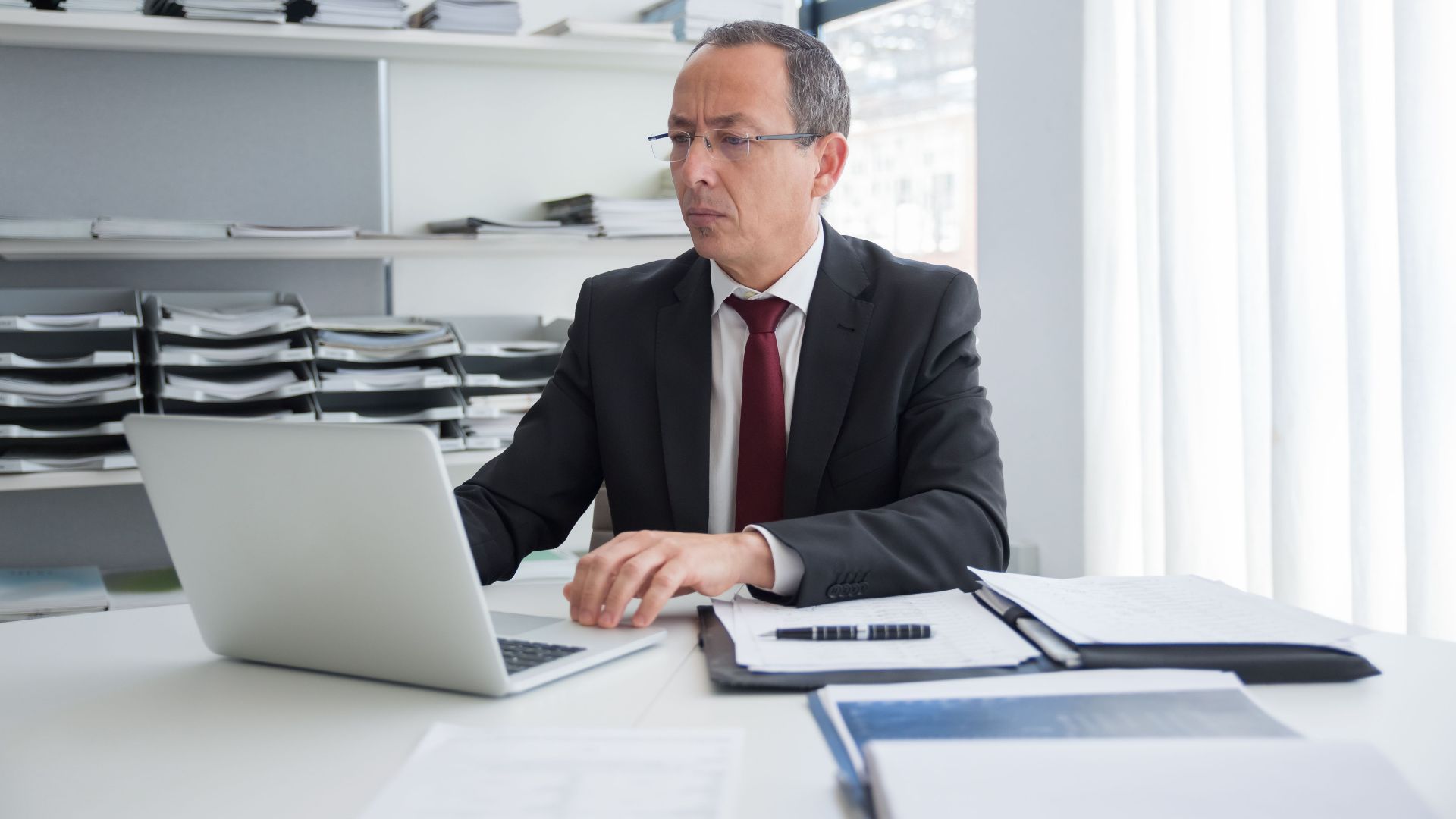 Focused businessman in a suit working at his office desk with a laptop and documents.
