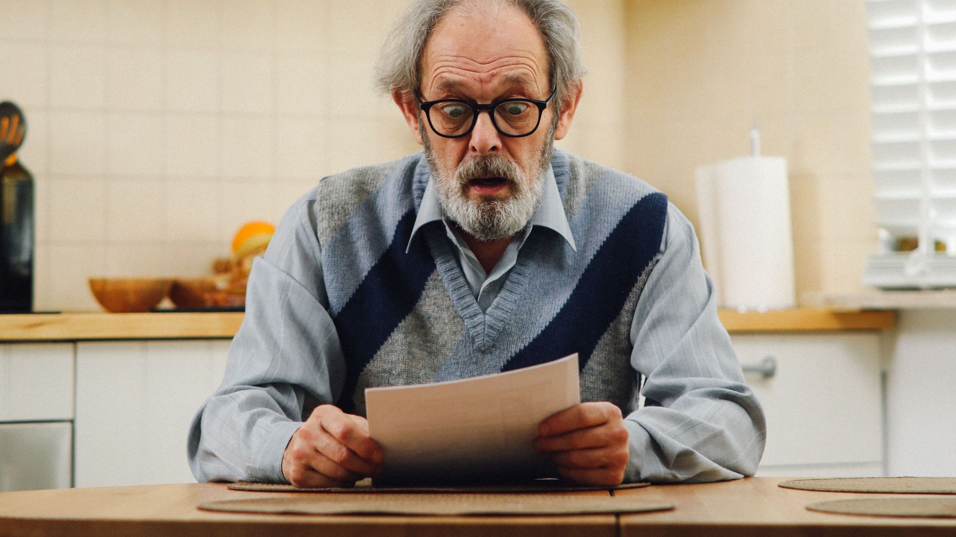 Senior man with eyeglasses reading a document at kitchen table, reflecting surprise.