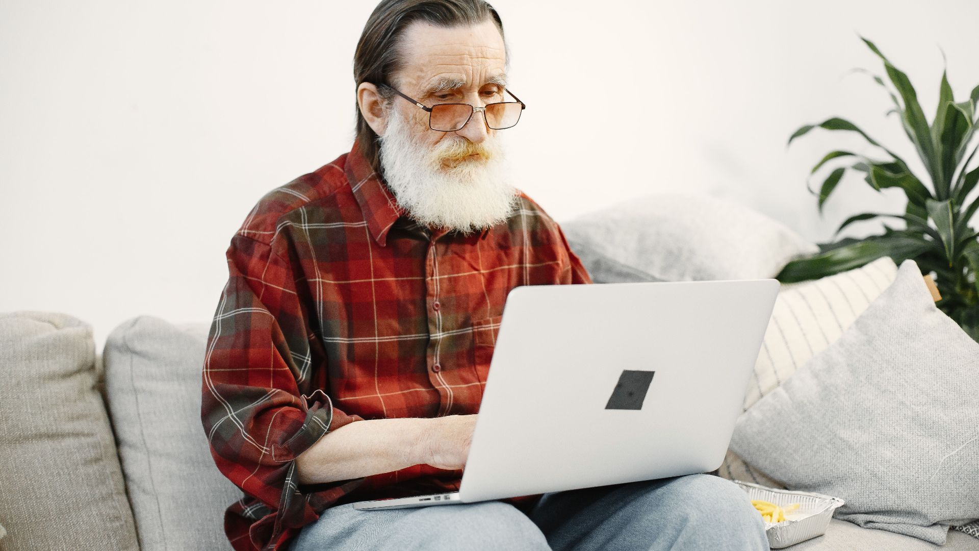An elderly man using a laptop on a comfortable sofa in a home setting.