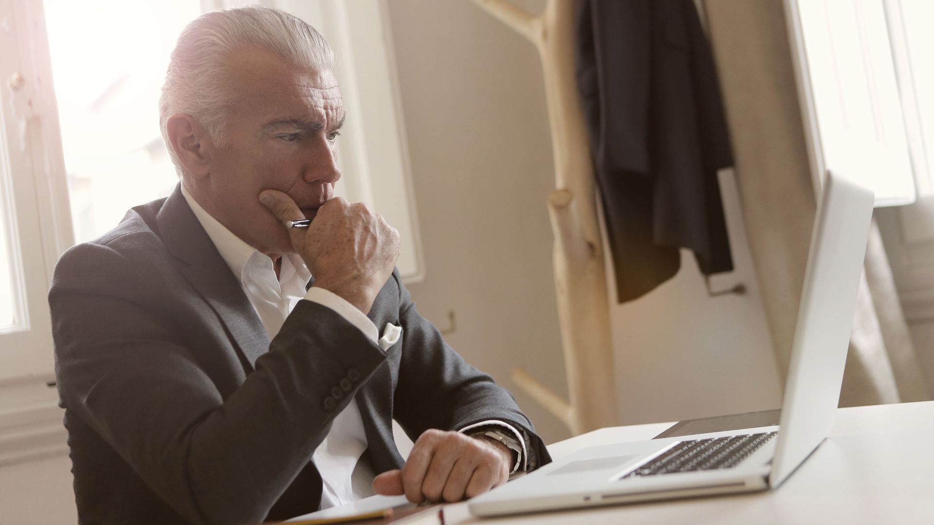 Elderly businessman in a suit thoughtfully working on a laptop at his desk.