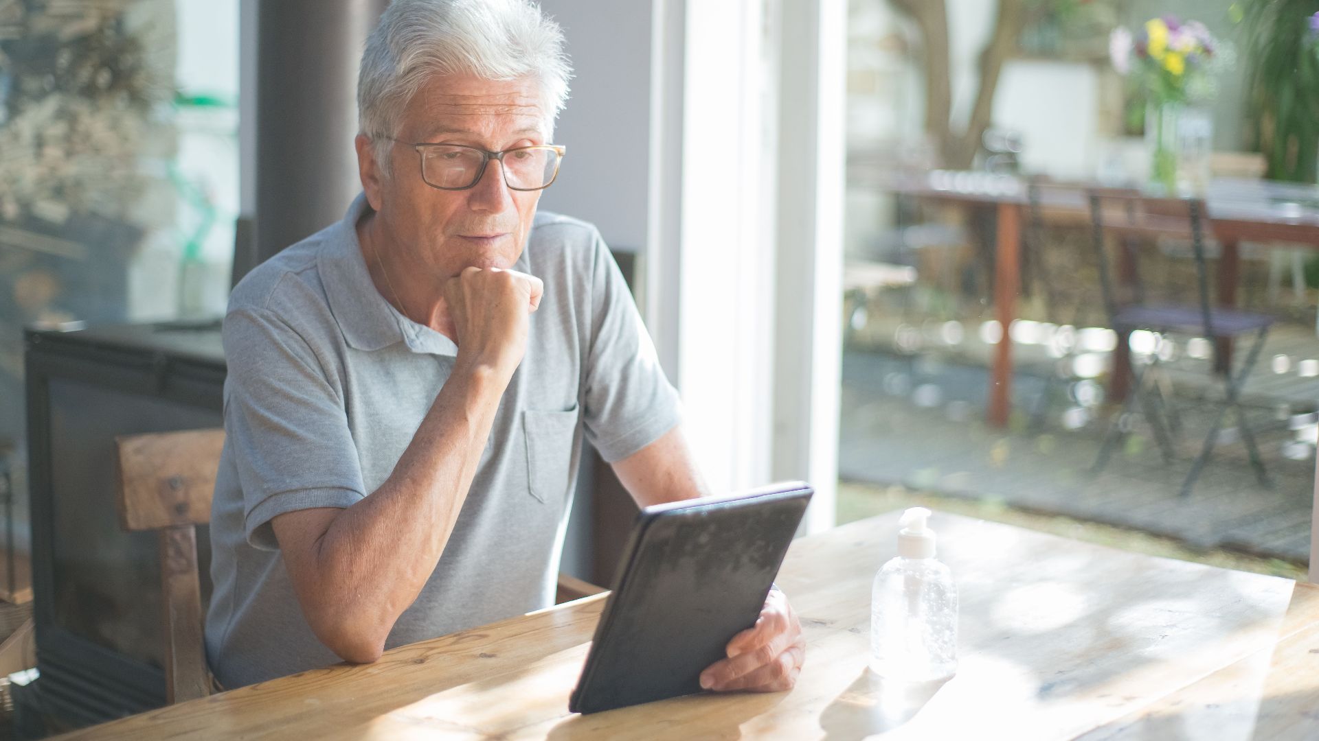 Senior man with eyeglasses reading a tablet at home in a sunlit room.