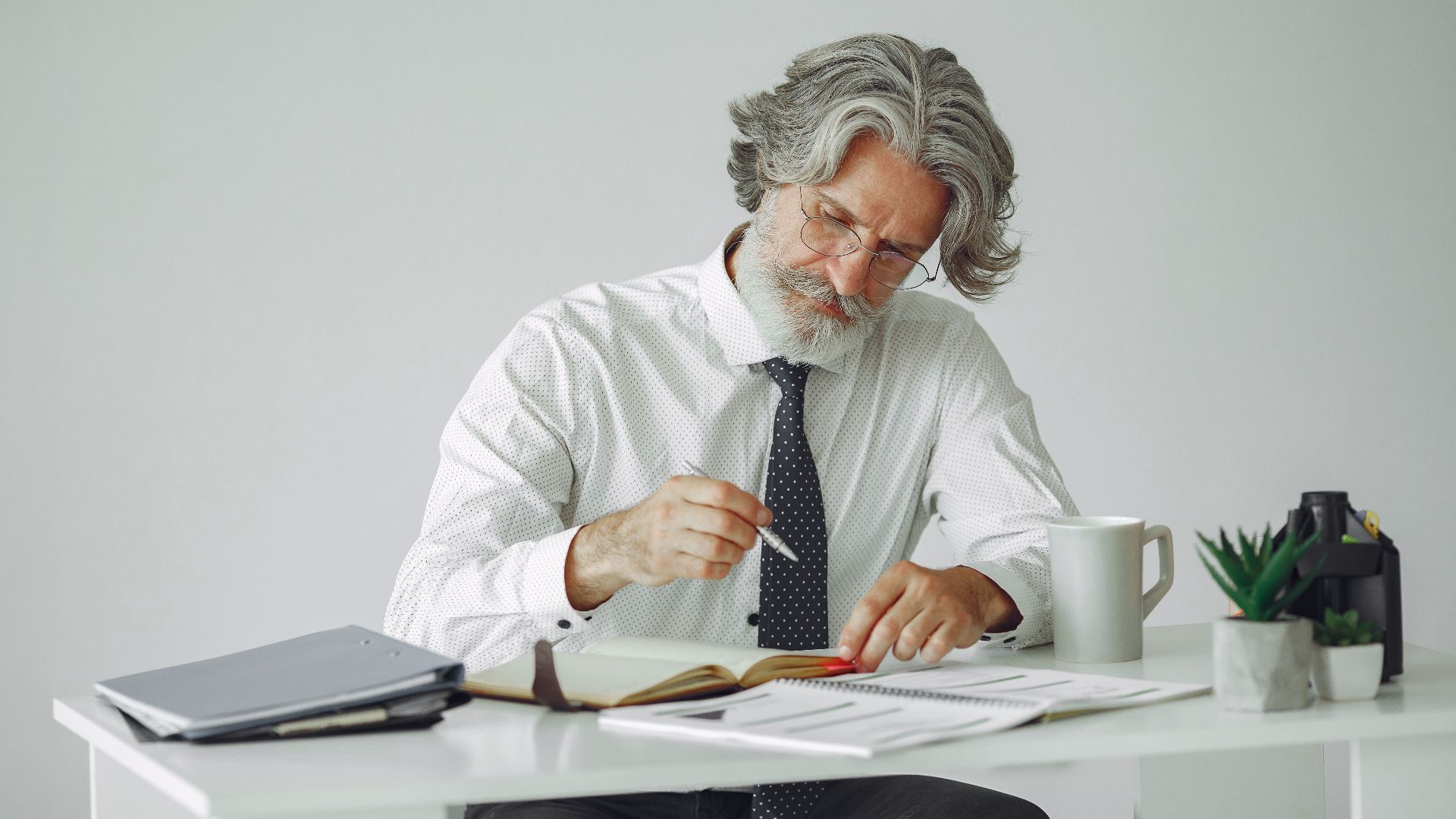 Senior man with grey hair working at desk with documents and coffee mug in office setting.