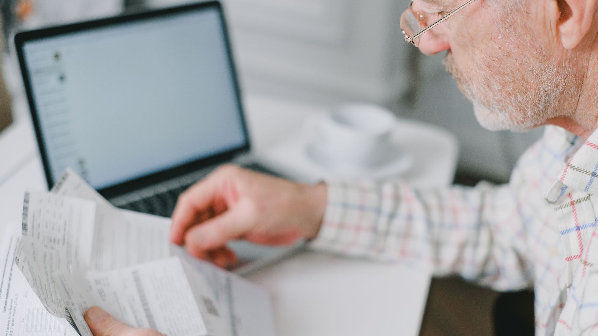 Elderly man wearing eyeglasses reading documents next to a laptop at home.