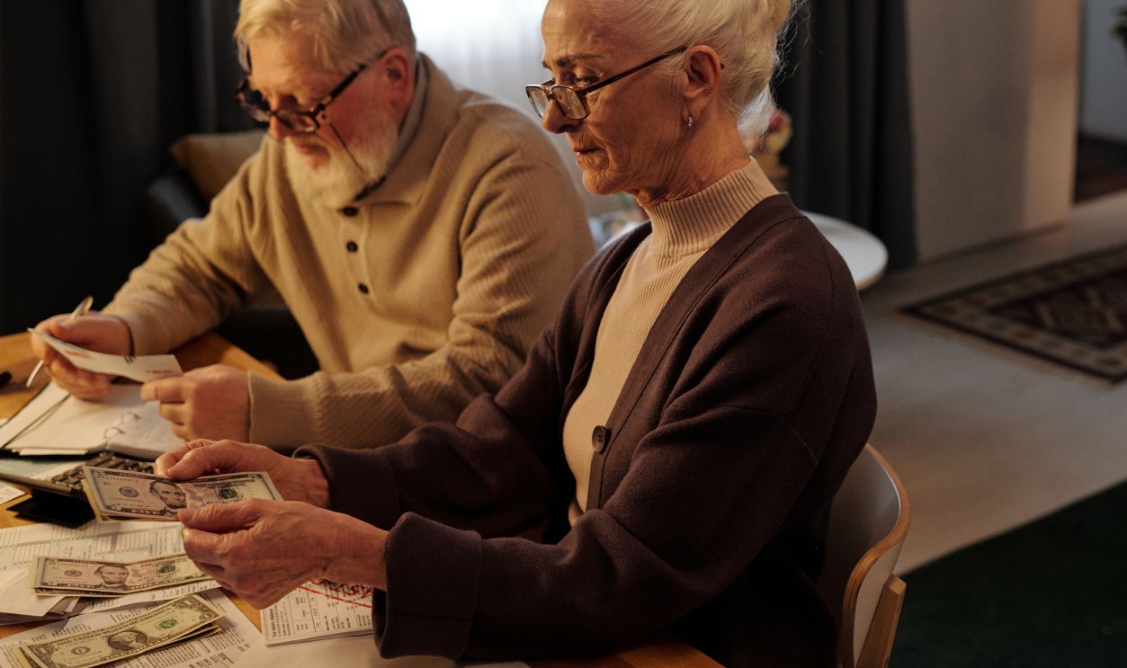 Senior woman in eyeglasses and cardigan sitting by table in spacious living room and counting money while her husband reading document