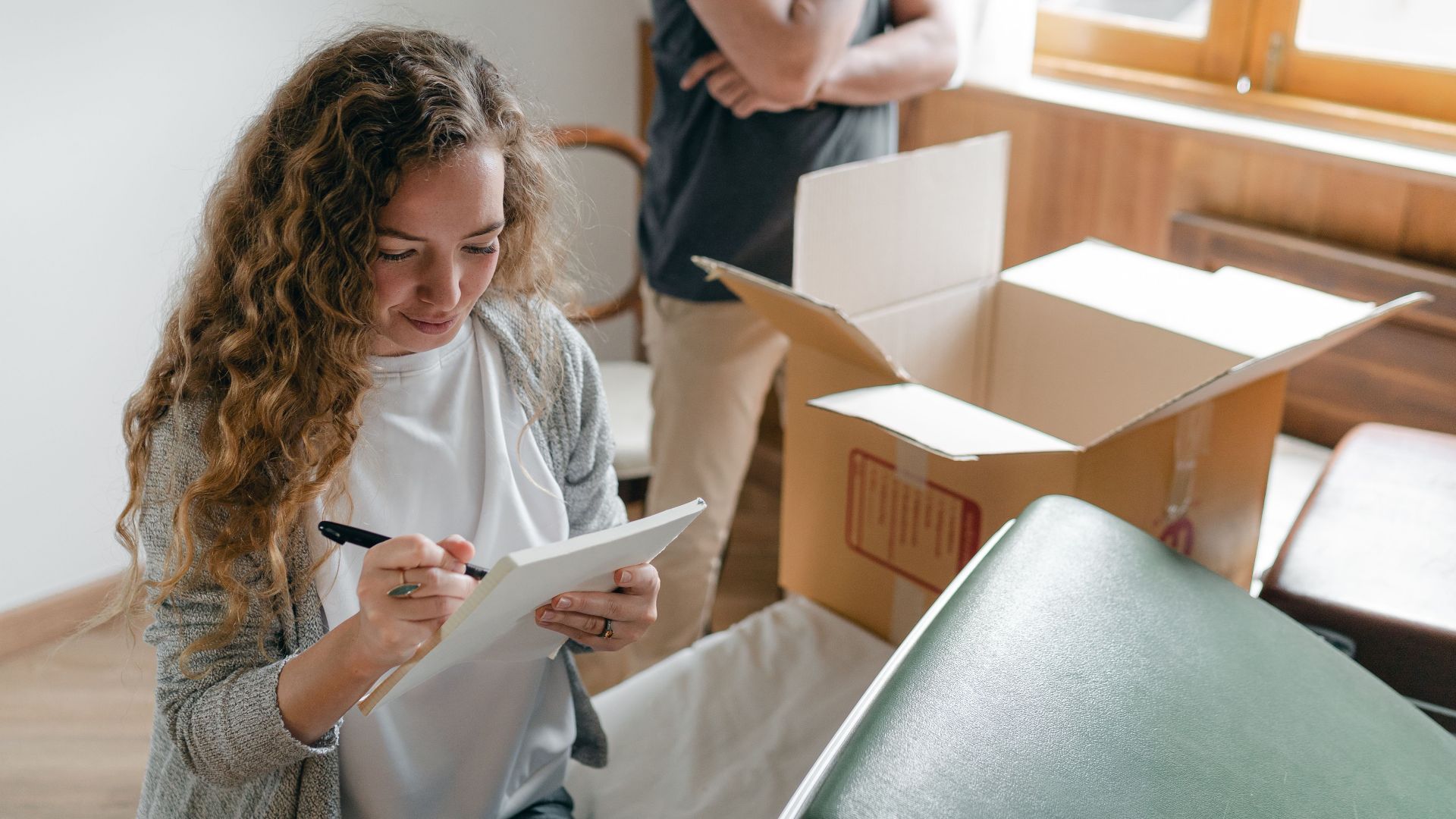 Pensive couple taking notes in bedroom near window while packing stuff in carton box to move into new apartment