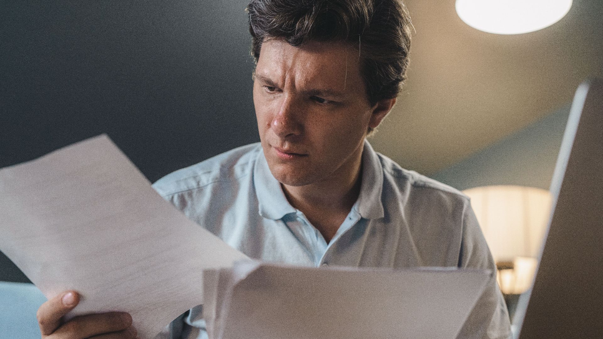 A man intently examines papers, seated indoors under warm lighting, focusing on his work.