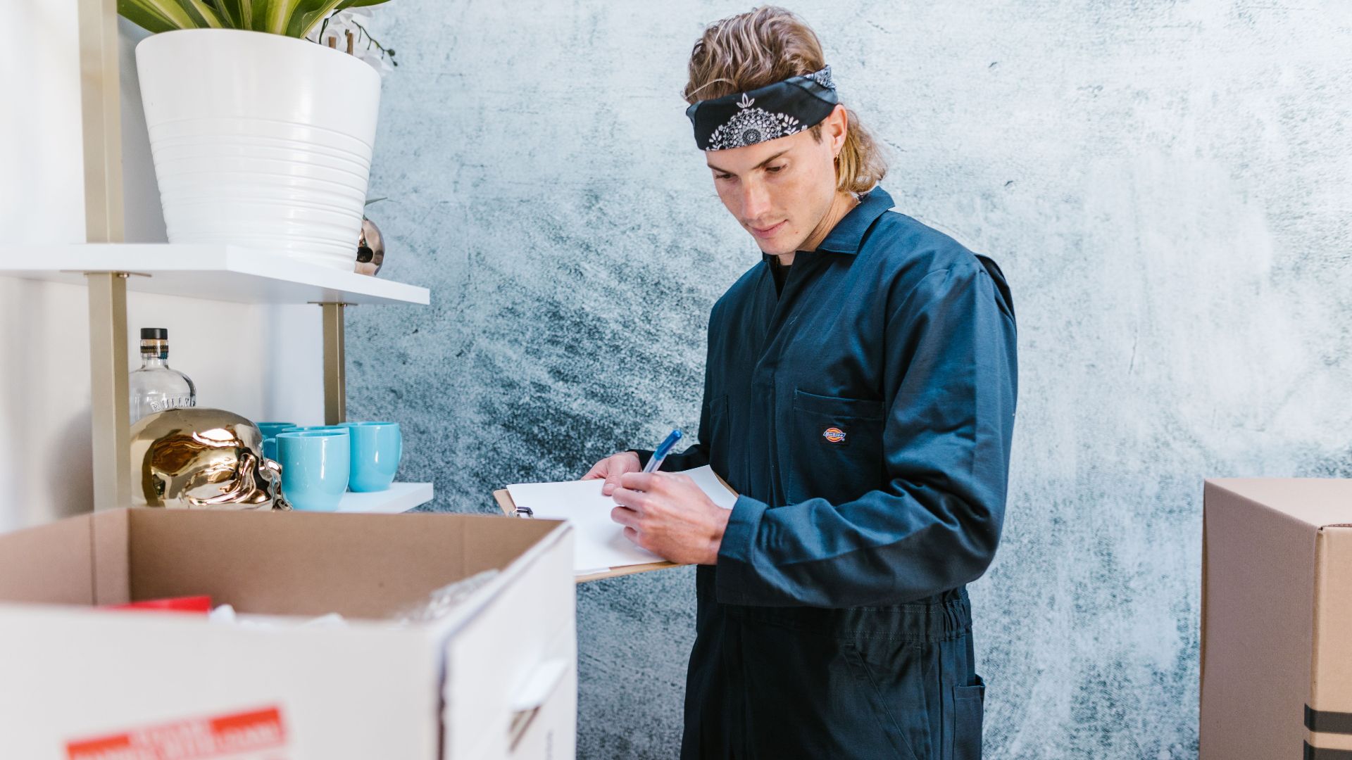 Man wearing overalls and bandana preparing for a home move, surrounded by boxes and fragile items.