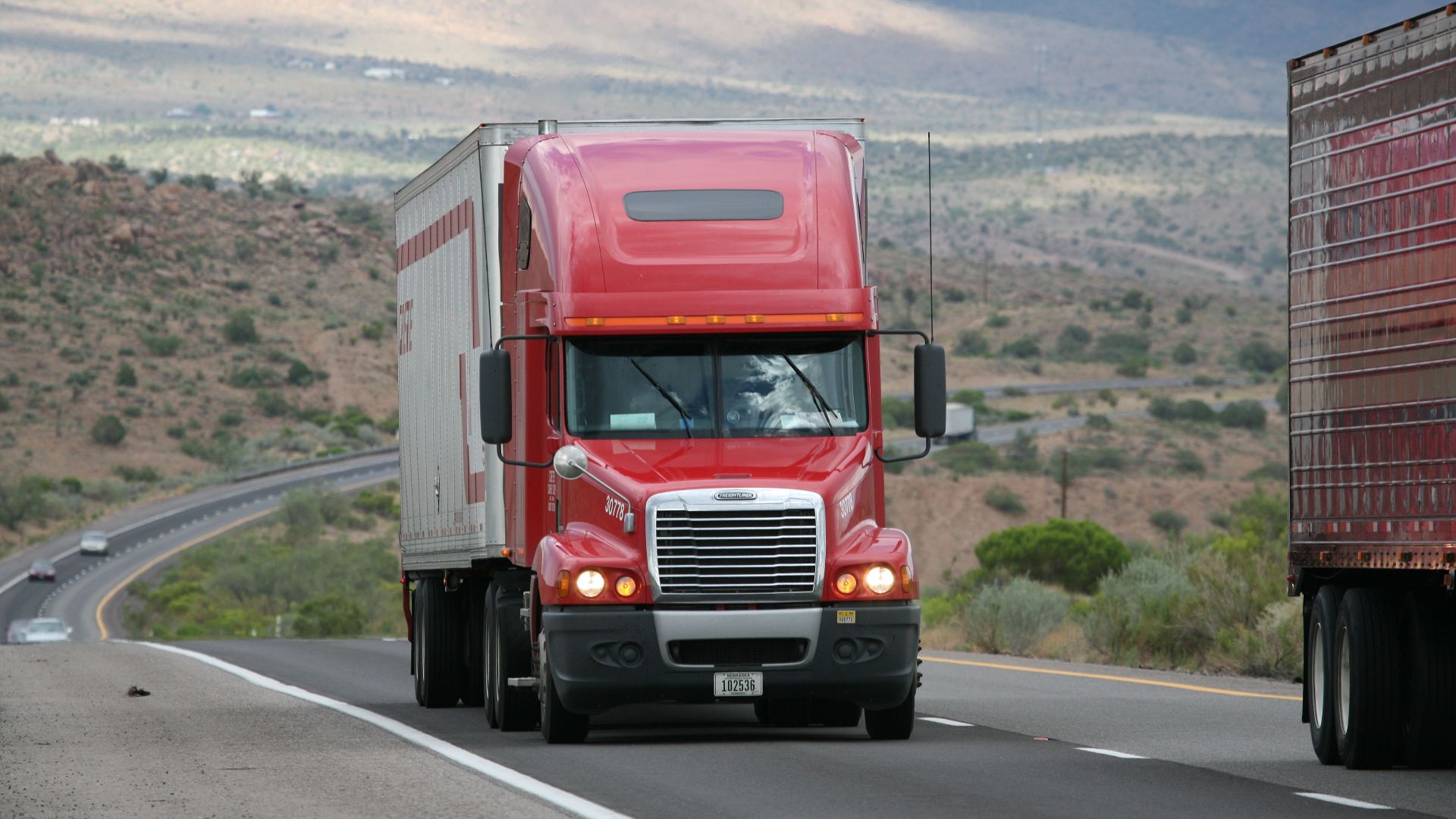 Red semi-truck driving through scenic highway in a mountainous landscape.