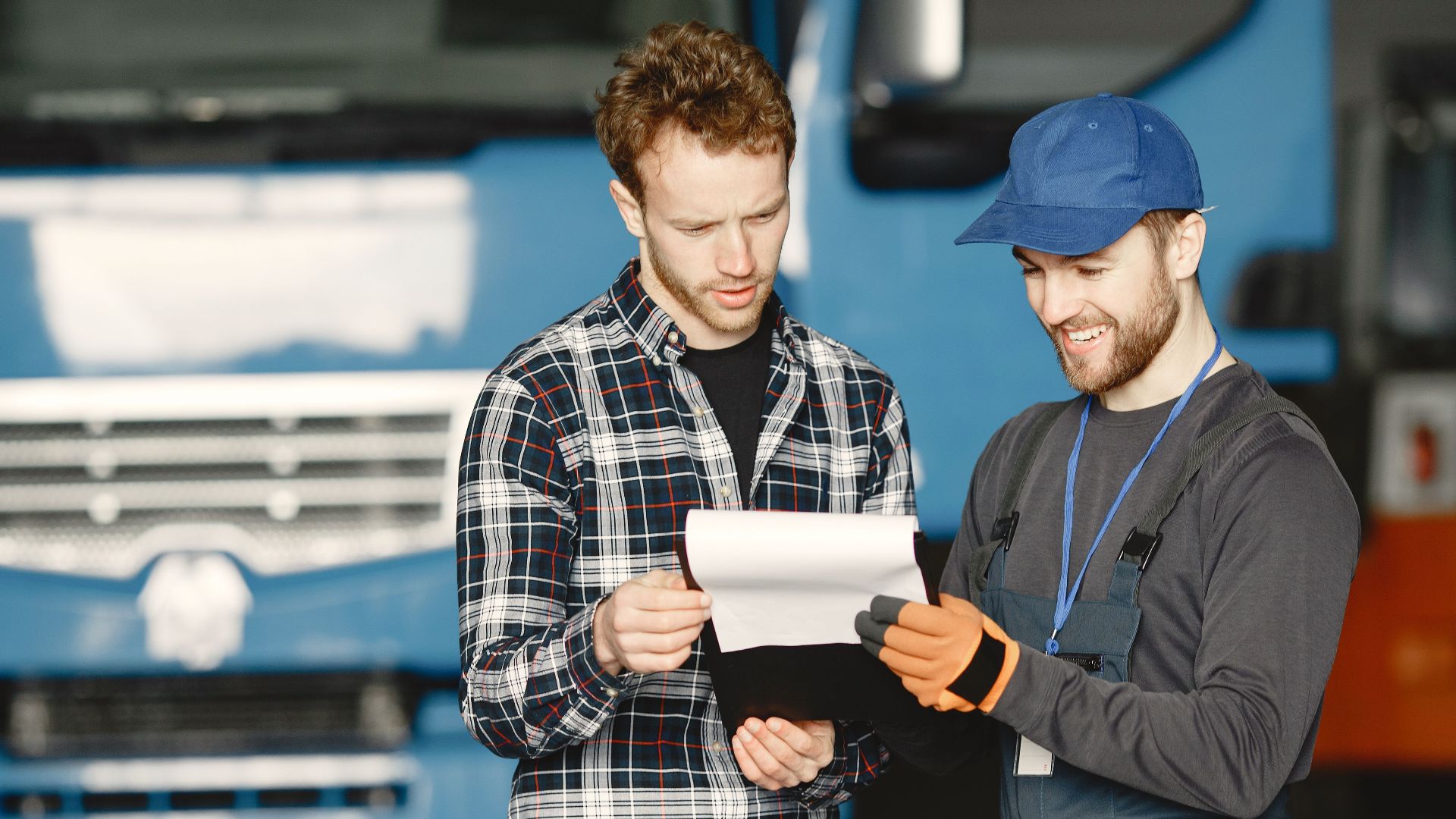 Two men in discussion over vehicle maintenance documents in a garage setting with trucks in the background.