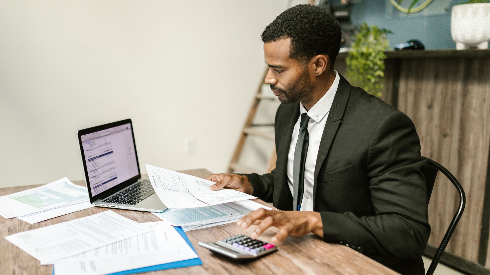 A businessman in a suit working on documents and using a laptop at an office desk.