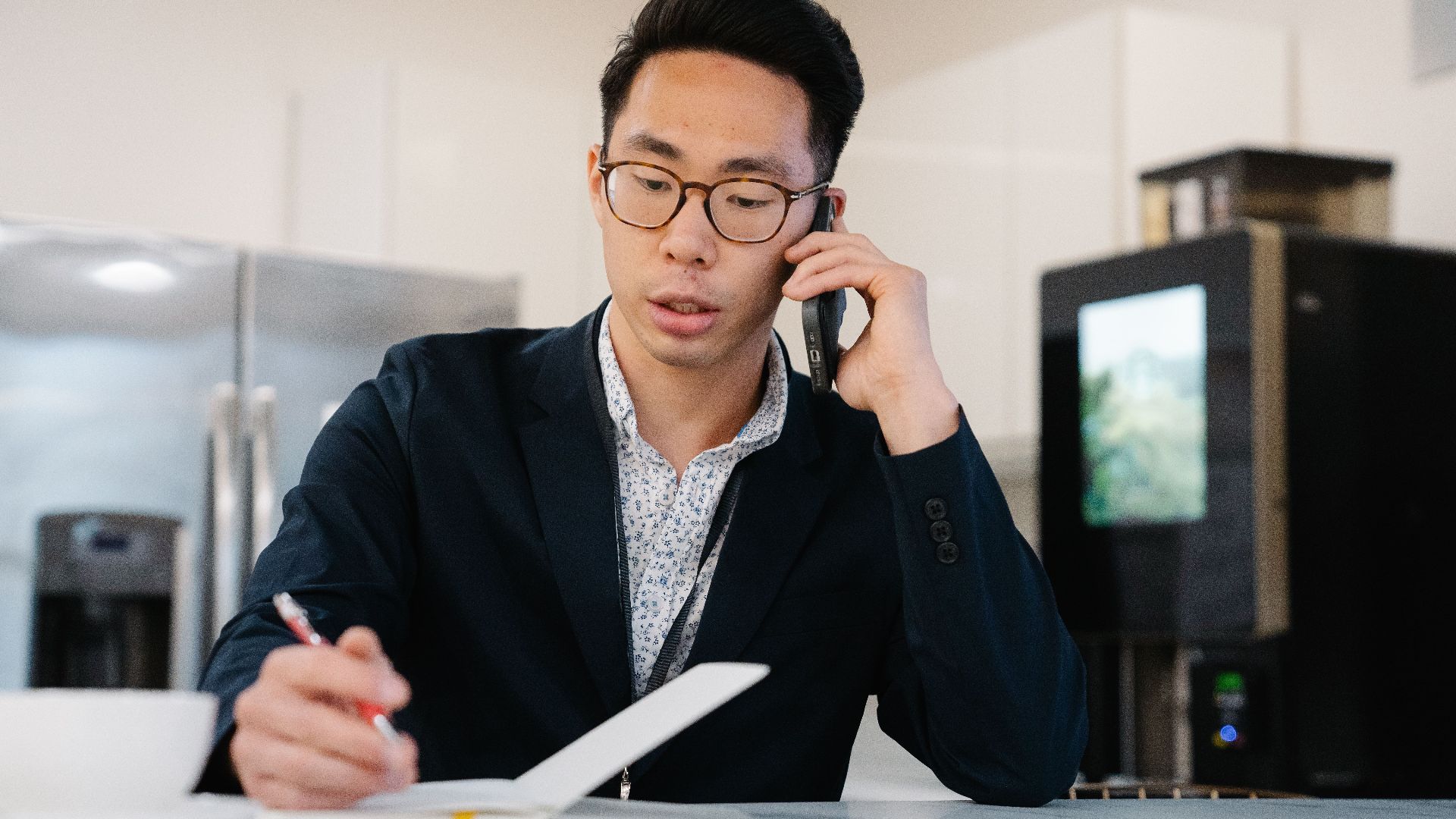Asian man in a suit jacket on a phone call, taking notes in an office.