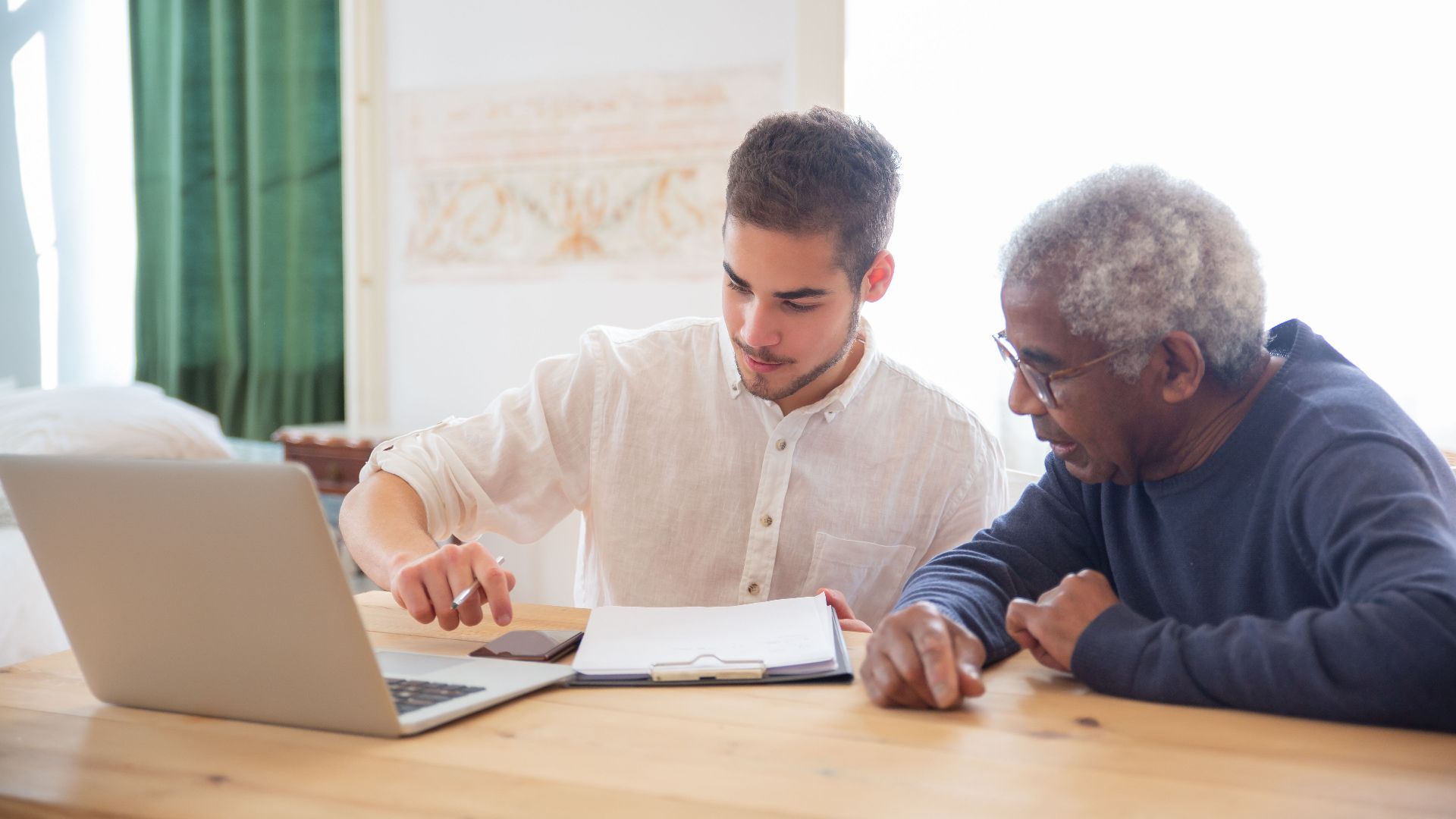 Young man teaches elderly gentleman to use technology at home.