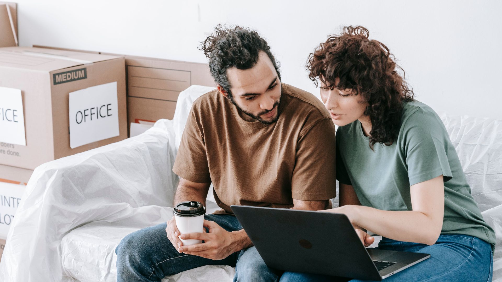 Young couple sits on a sofa using a laptop during a move, surrounded by boxes and sipping coffee.