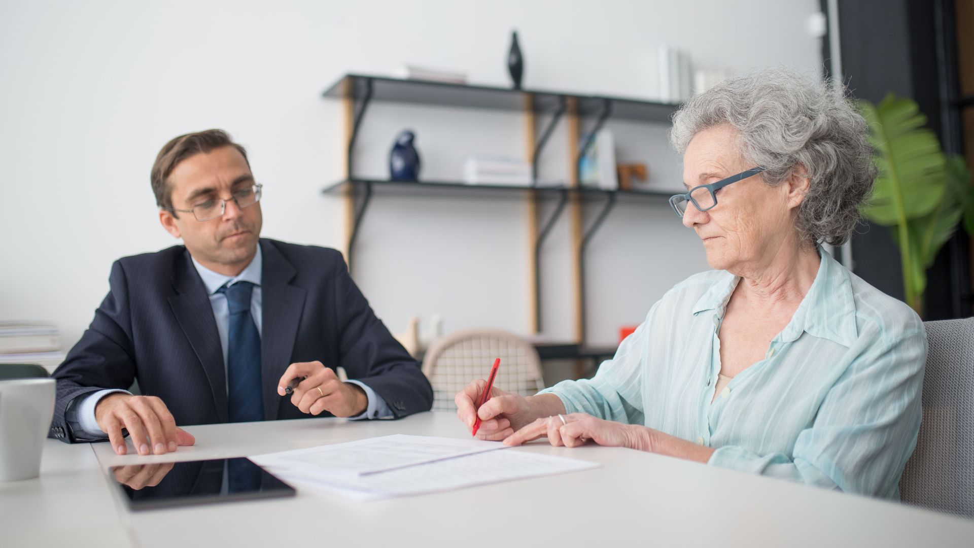 Elderly woman signing paperwork in modern office with consultant.