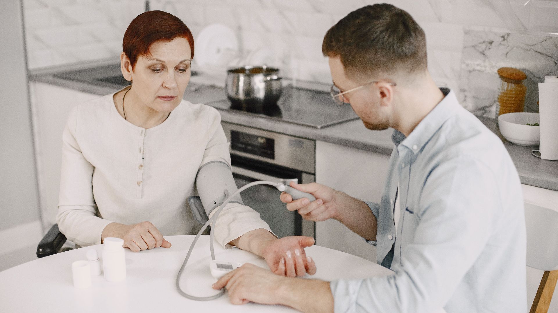 Adult man monitoring elderly woman's blood pressure at kitchen table using sphygmomanometer.