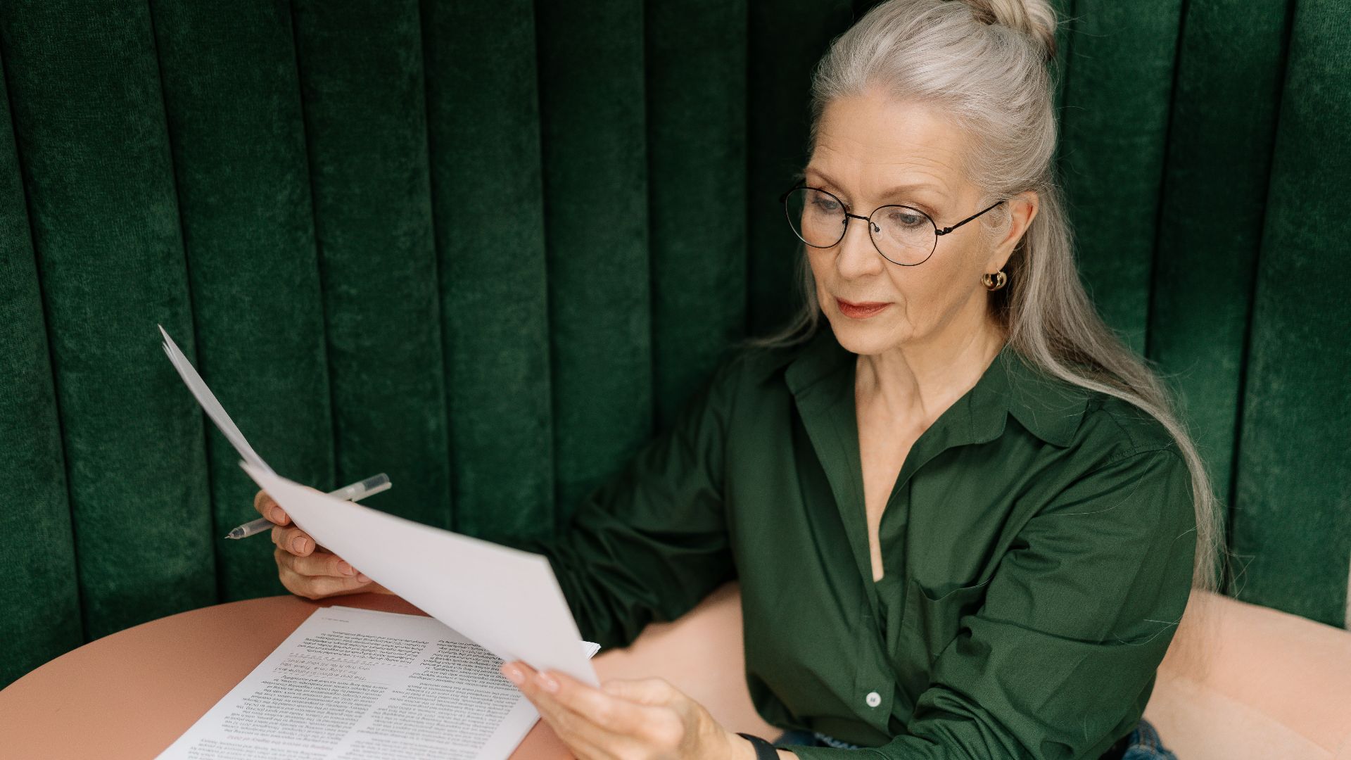 Senior woman wearing glasses, reading documents at a desk indoors, showcasing professional focus.
