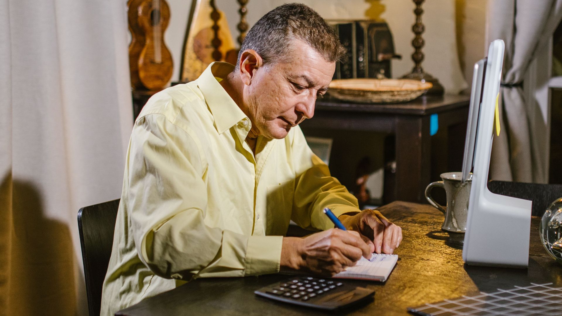 Senior man wearing yellow shirt writing notes at desk with lamp, calculator, and glass sphere nearby.
