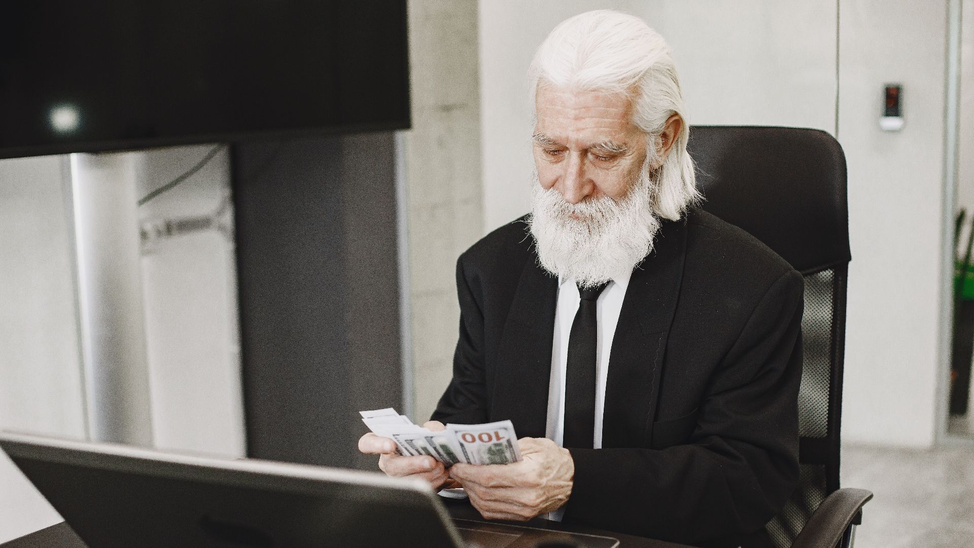 Senior businessman in suit counting money at office desk with a laptop. Elegance and financial focus in modern workspace.