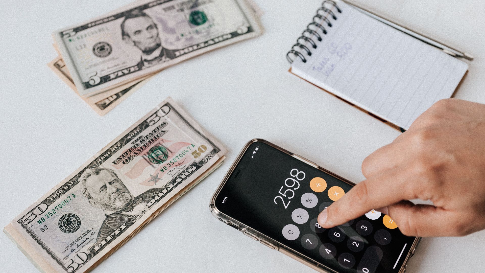 From above of crop anonymous economist calculating total amount of income using calculator app on cellphone near piles of greenbacks and notebook with pen