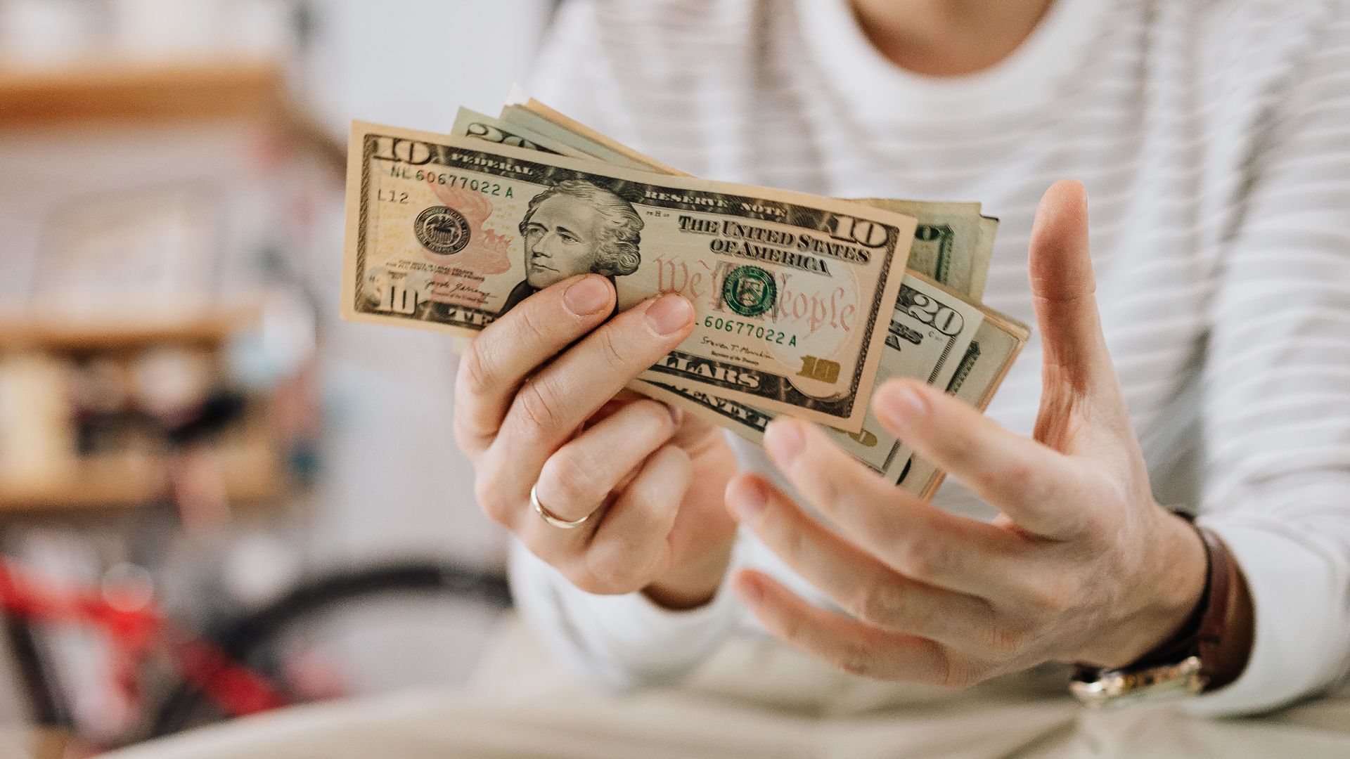 Man wearing casual clothes counts dollar bills while sitting on a sofa indoors.