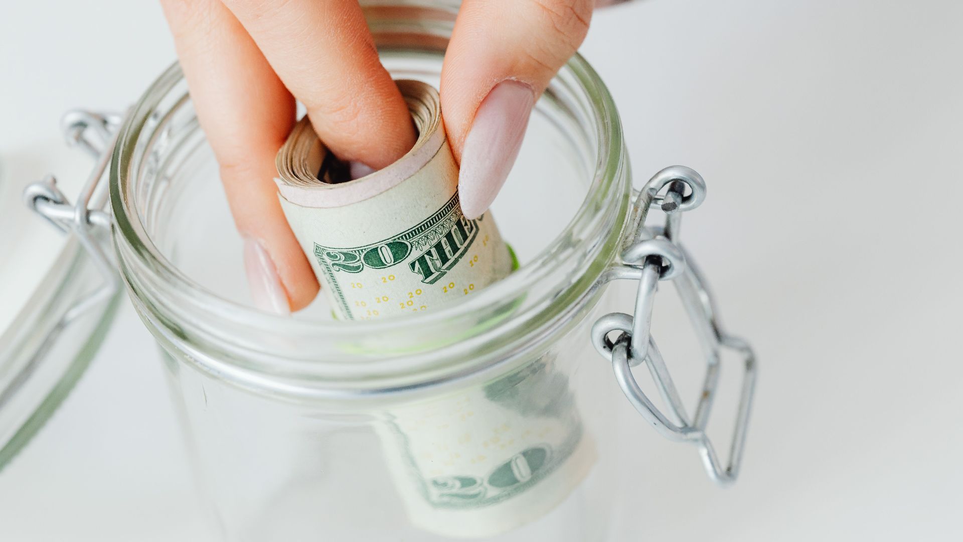 A close-up of a hand placing rolled dollars into a glass jar, symbolizing savings.