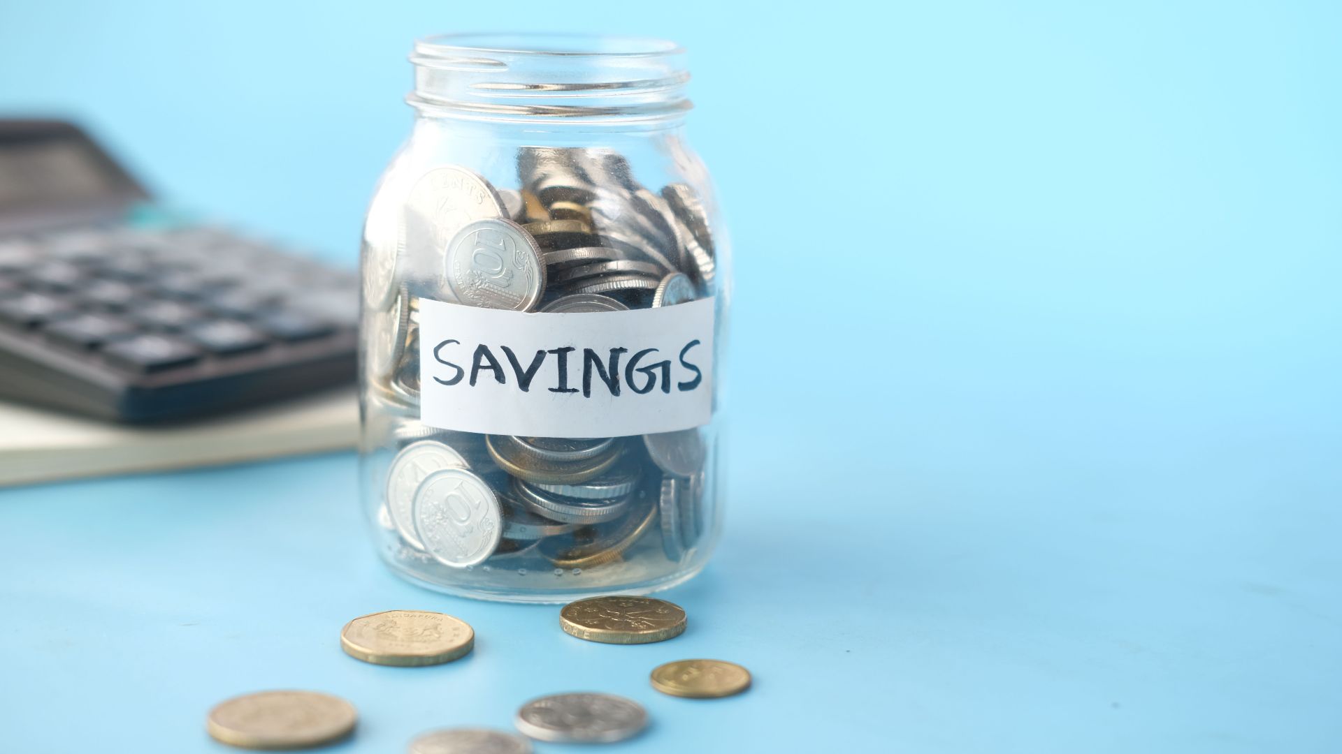 Glass jar labeled 'Savings' filled with coins, beside a calculator on a blue background.