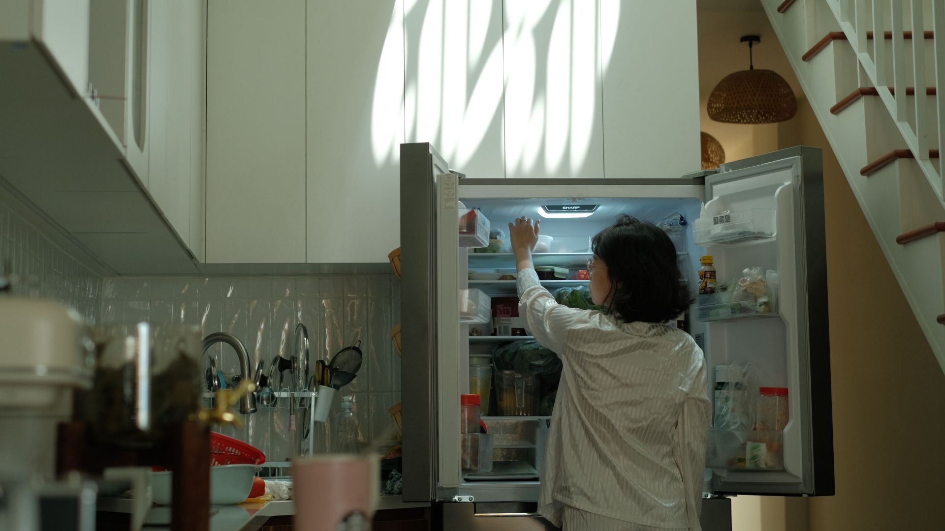 A woman in pajamas checks the refrigerator in a warmly lit cozy kitchen.