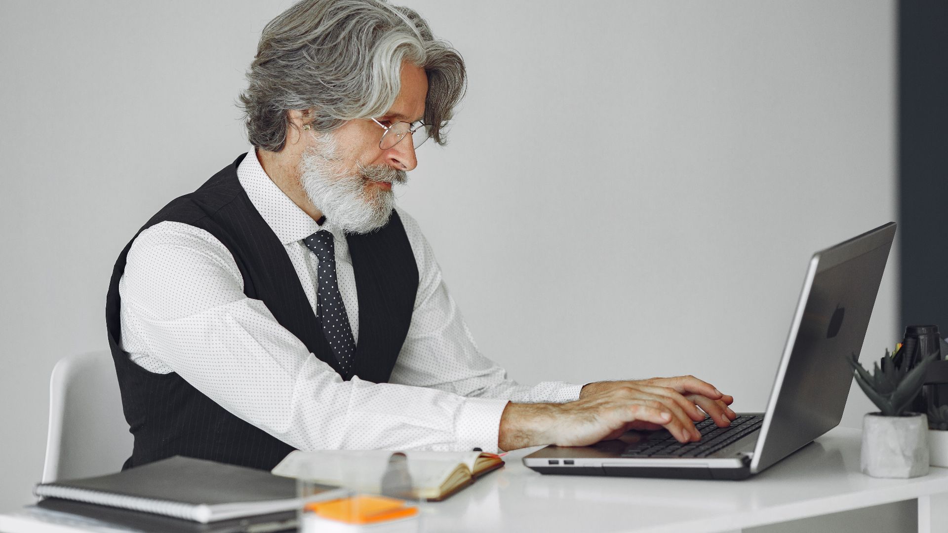 Elegant senior man in office attire typing on a laptop at a desk.