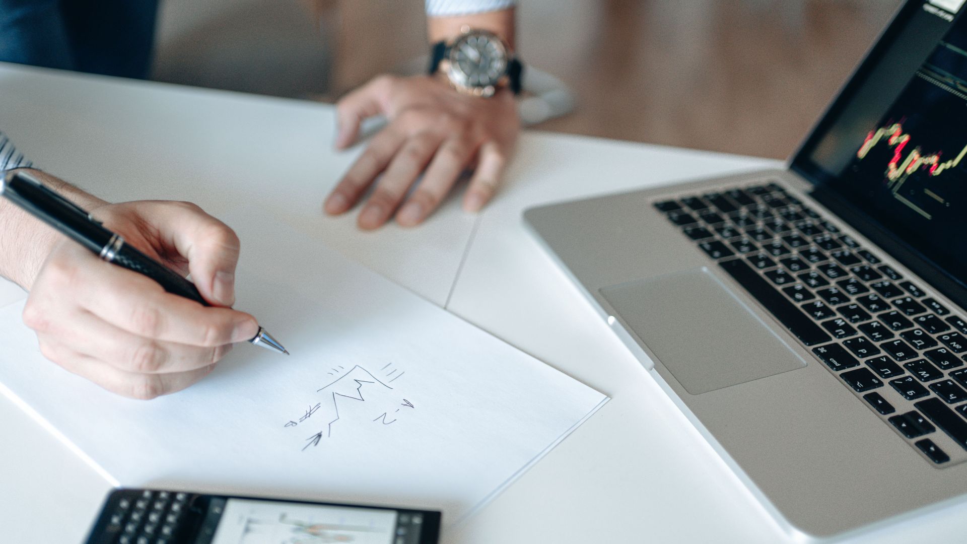 Close-up of hands drawing stock trends on paper with laptop and smartphone on desk.