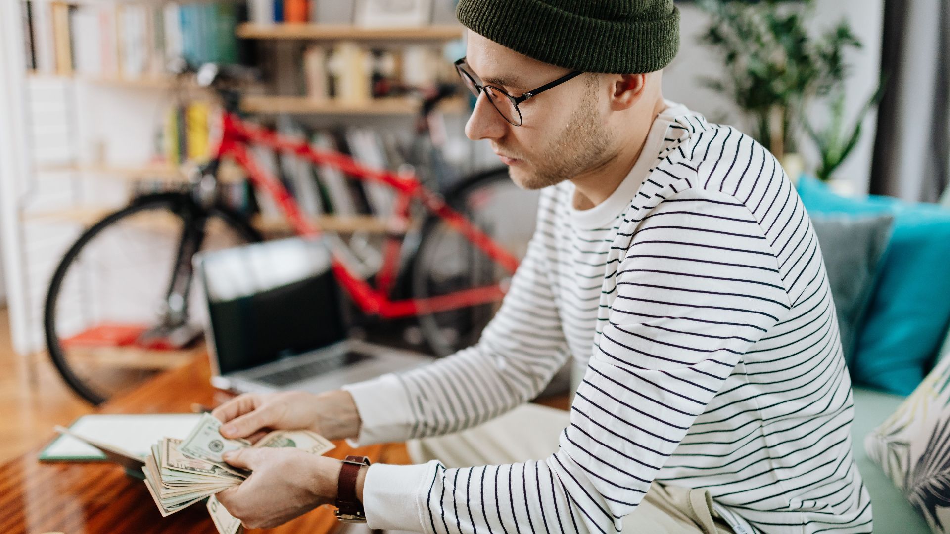 Man in striped shirt counting cash indoors, with a bicycle in the background.