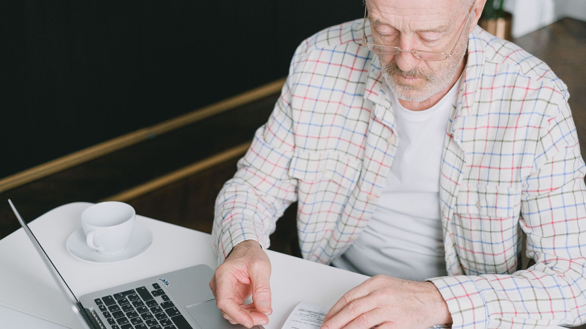 Elderly man with eyeglasses reviewing documents at a laptop. Indoor setting with natural light.