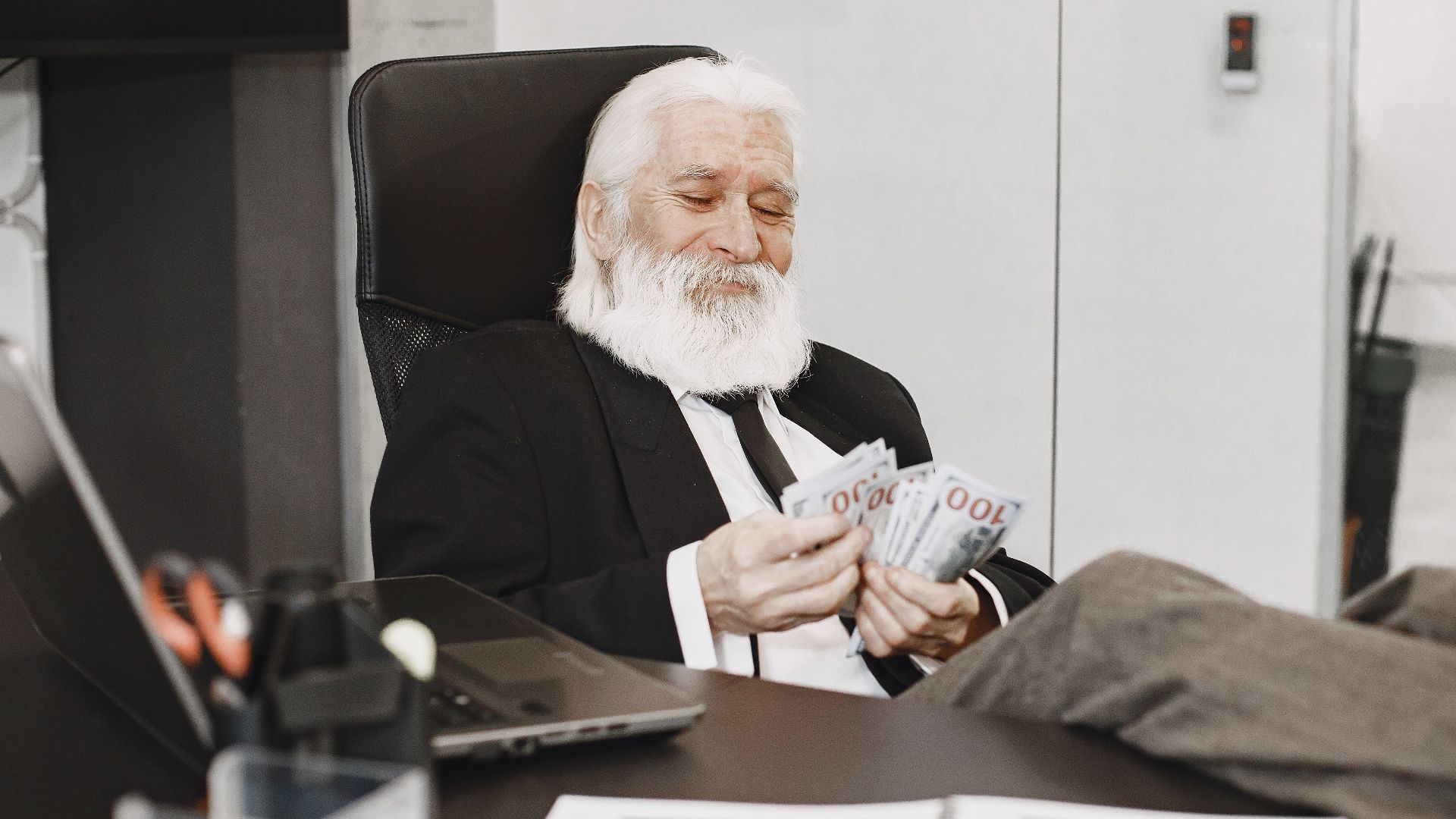 Elderly businessman relaxing at desk, counting money notes in a modern office setting.