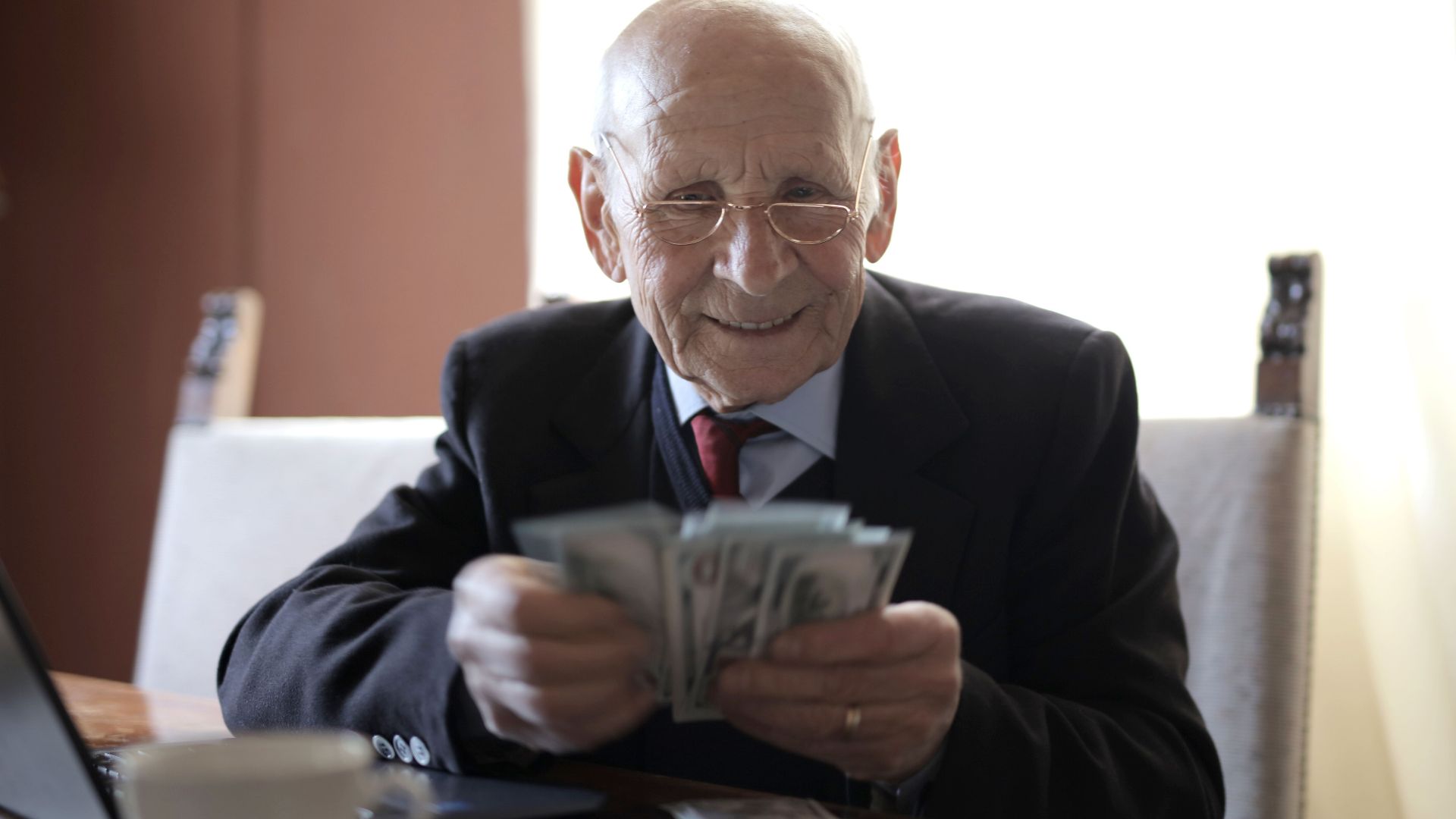 Positive senior businessman in formal suit and eyeglasses counting money bills while sitting at wooden table with cup of beverage and near opened laptop