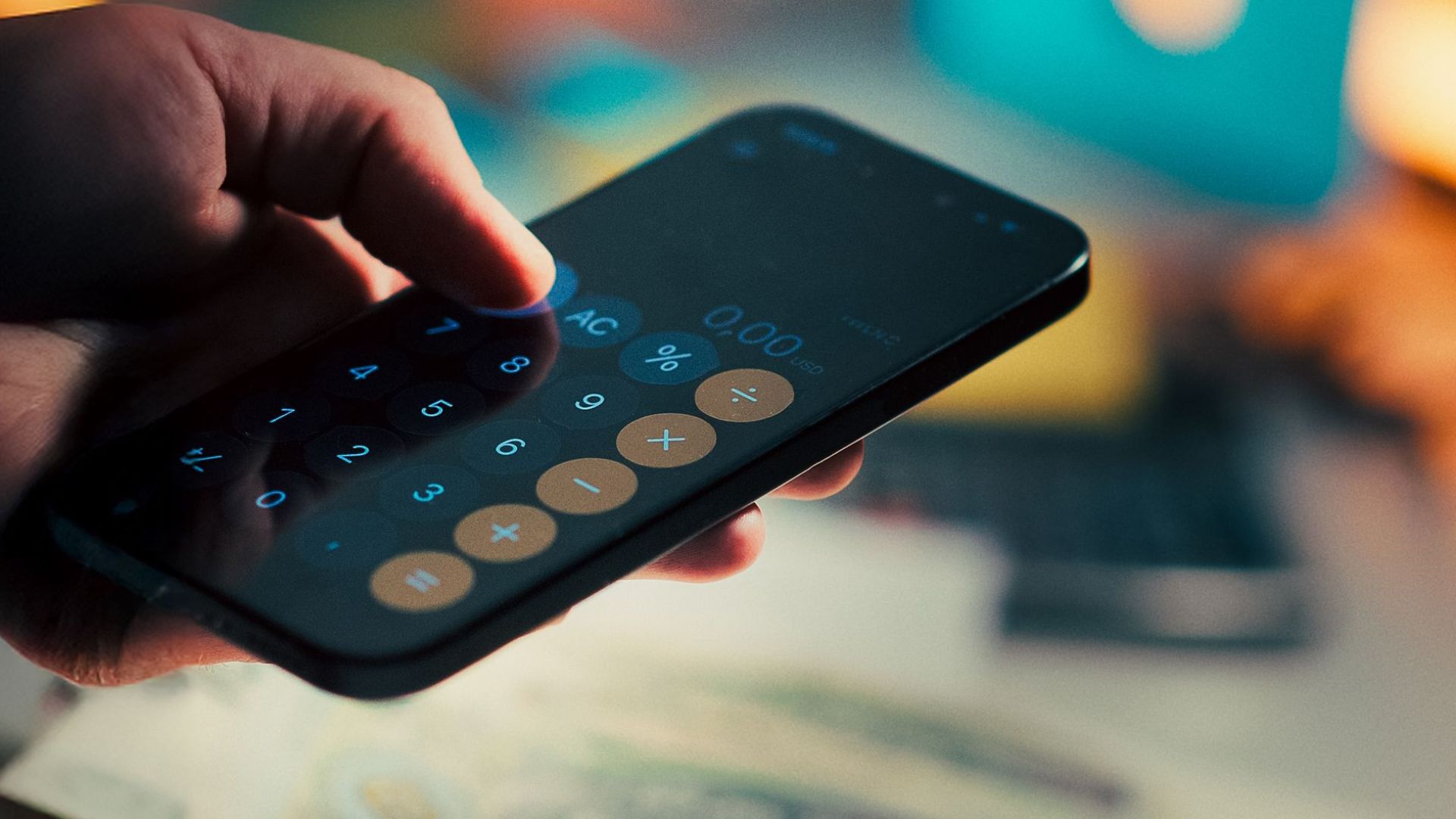 A hand holds a smartphone running a calculator app, with cash and a notebook on a desk, symbolizing budgeting and finance management.