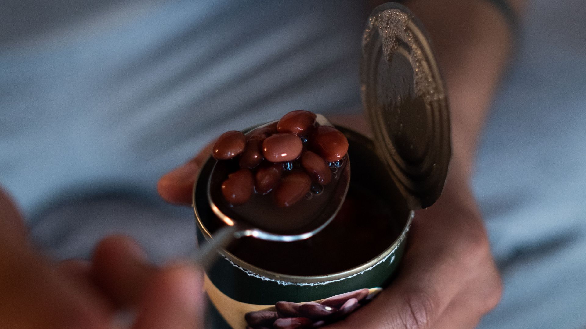 A person holding a spoonful of beans from an open can, focusing on details and texture.