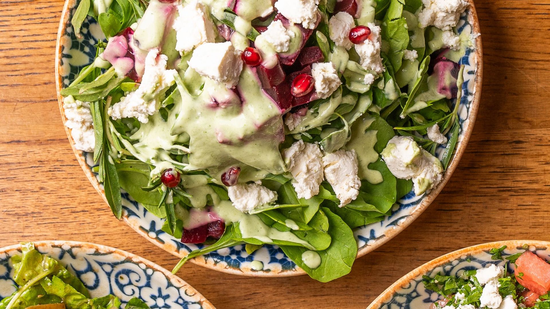 Top view of three fresh salads with feta cheese, vegetables, and herbs on a wooden table.