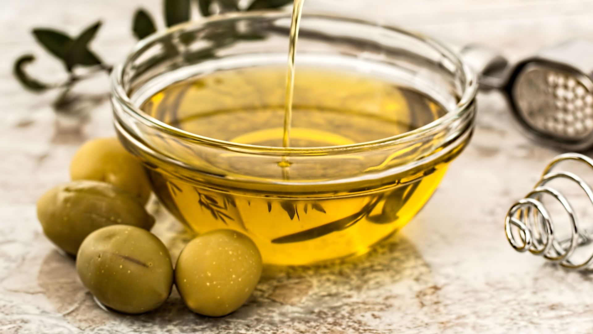 Close-up of olive oil being poured into a glass bowl surrounded by fresh olives and kitchen tools.