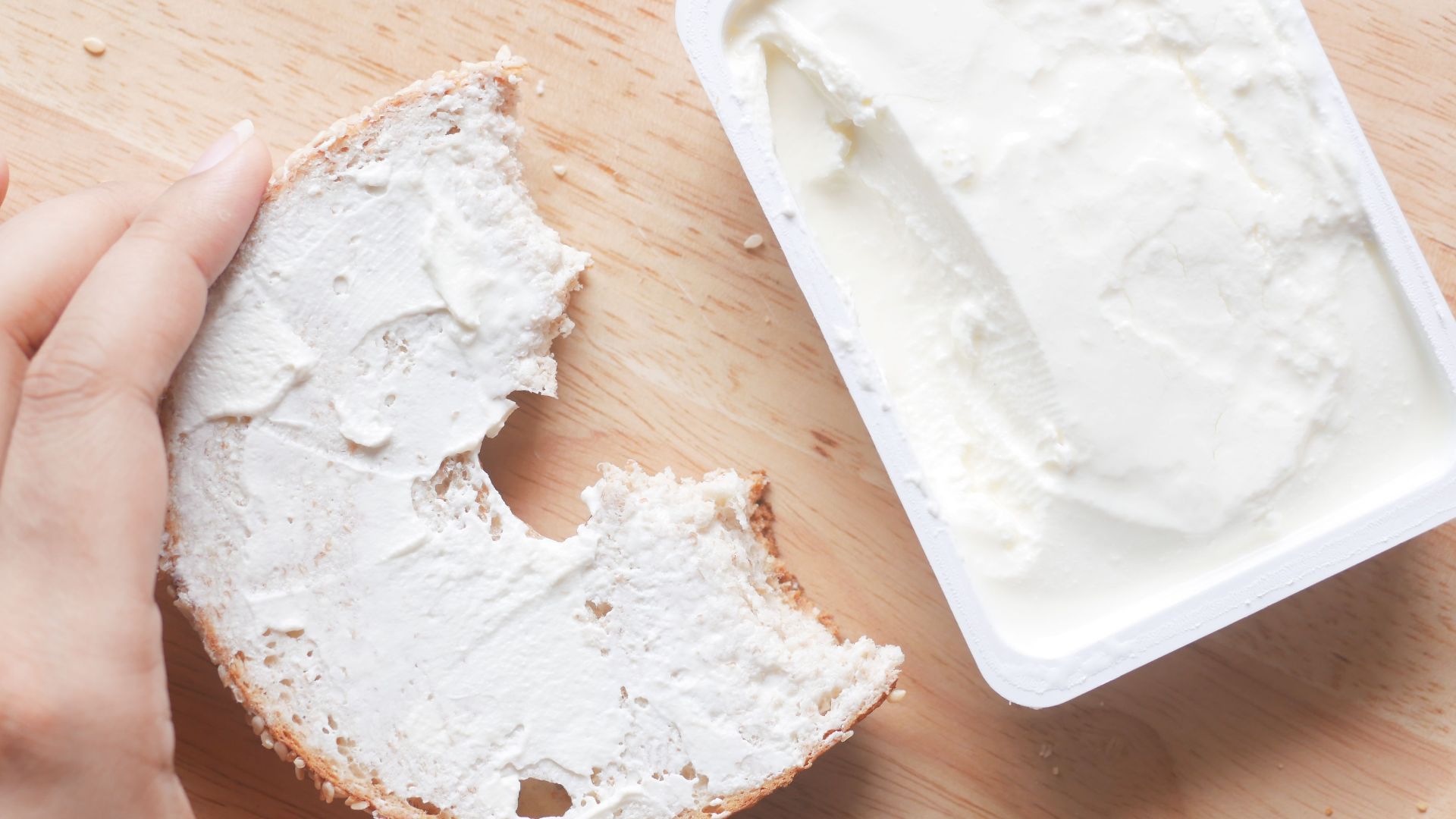 Close-up of a hand holding a bagel with creamy cheese spread next to an open container.