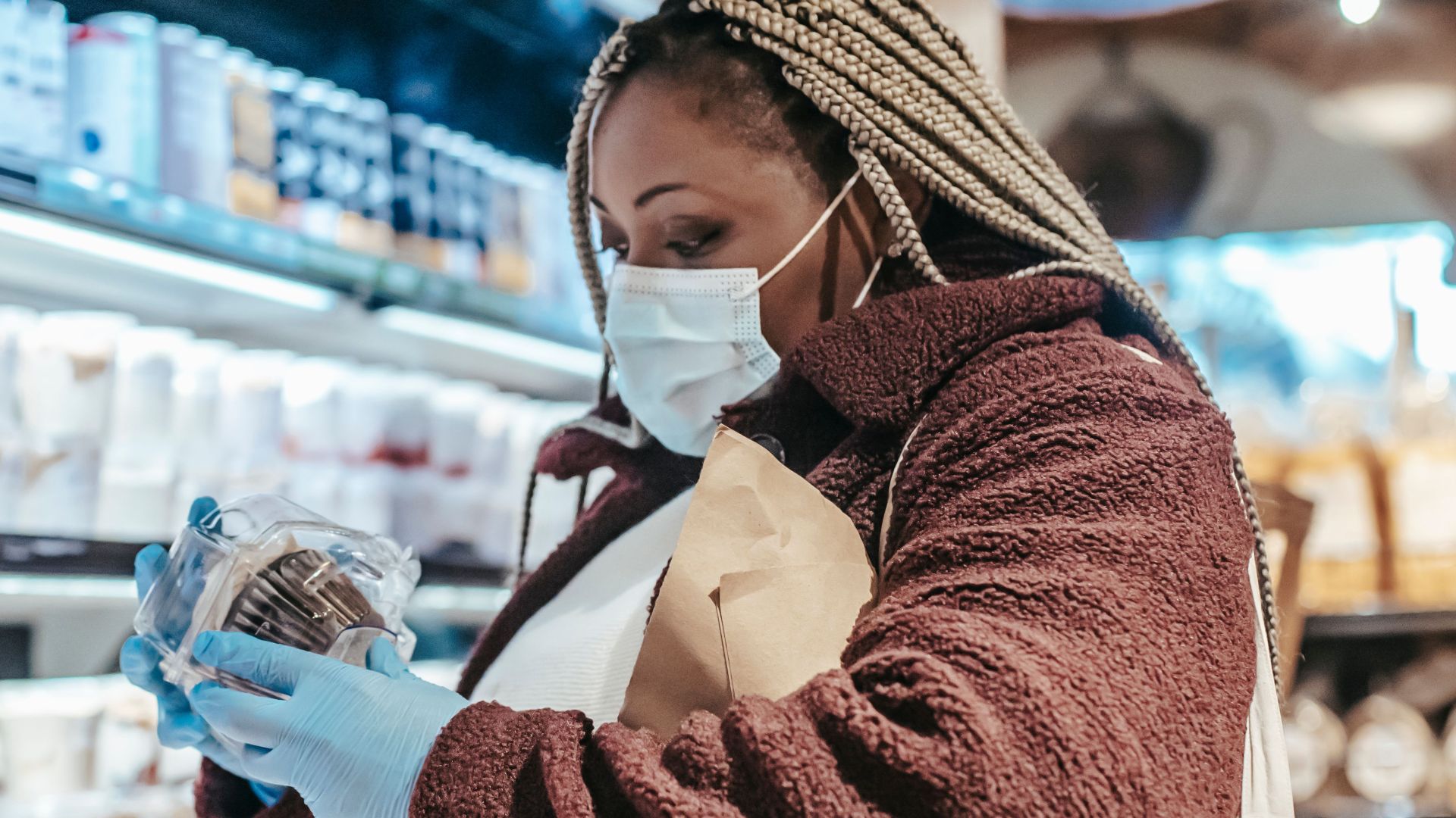 Side view of African American woman in protective gloves and mask reading sticker on product in grocery store