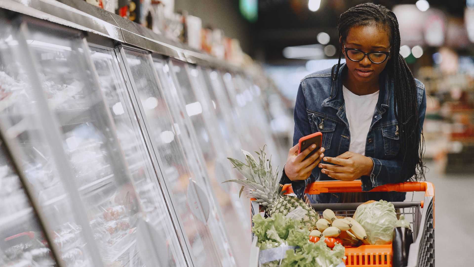 Woman in a grocery store using smartphone while pushing a shopping cart filled with groceries.