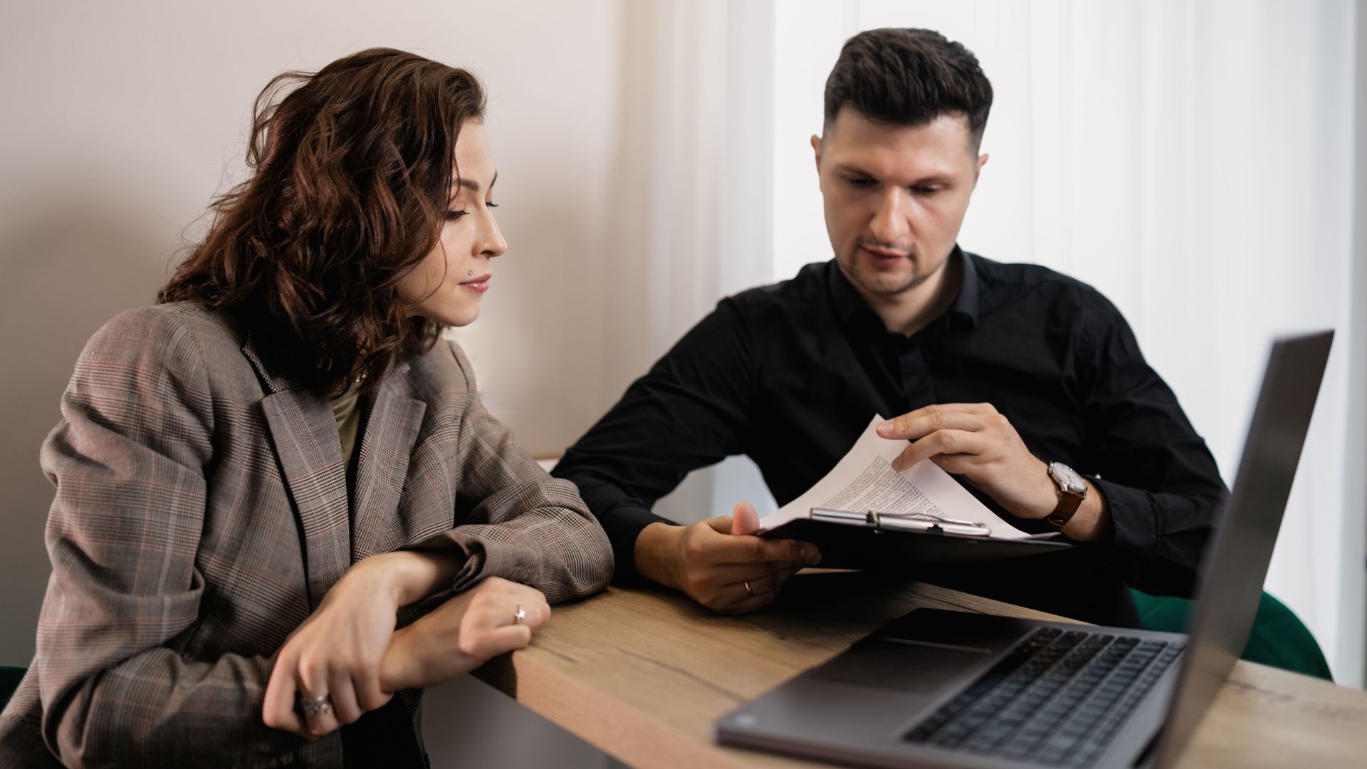 Two professionals discussing documents at a desk with a laptop, focused on collaboration.