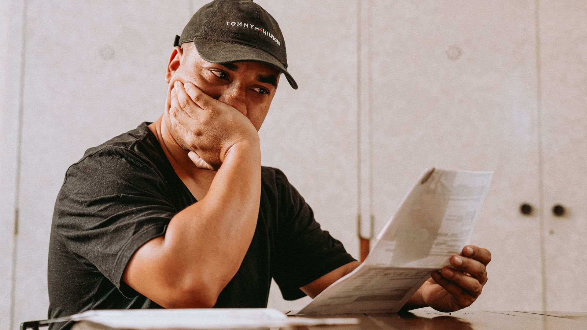 A man sits indoors, deeply focused on reviewing paperwork, expressing concern and contemplation.