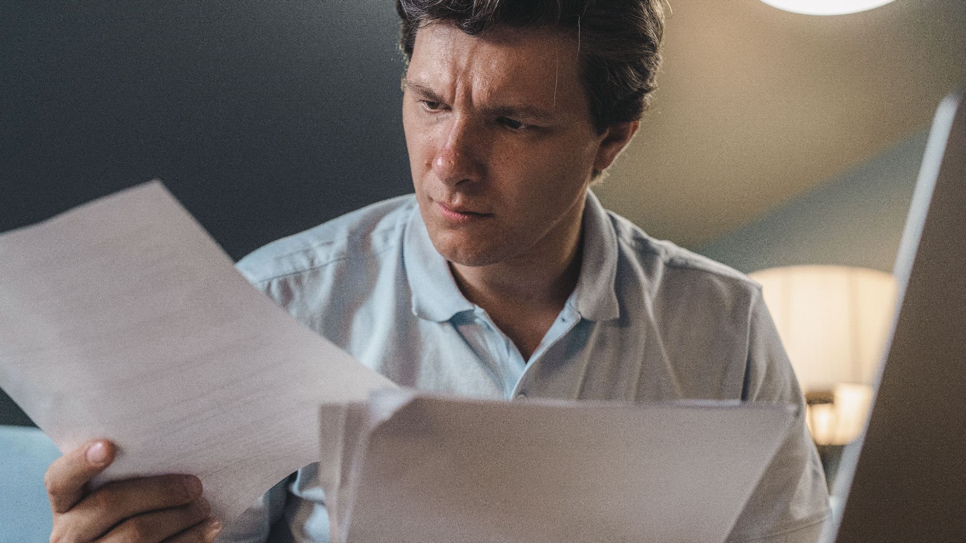 A man intently examines papers, seated indoors under warm lighting, focusing on his work.