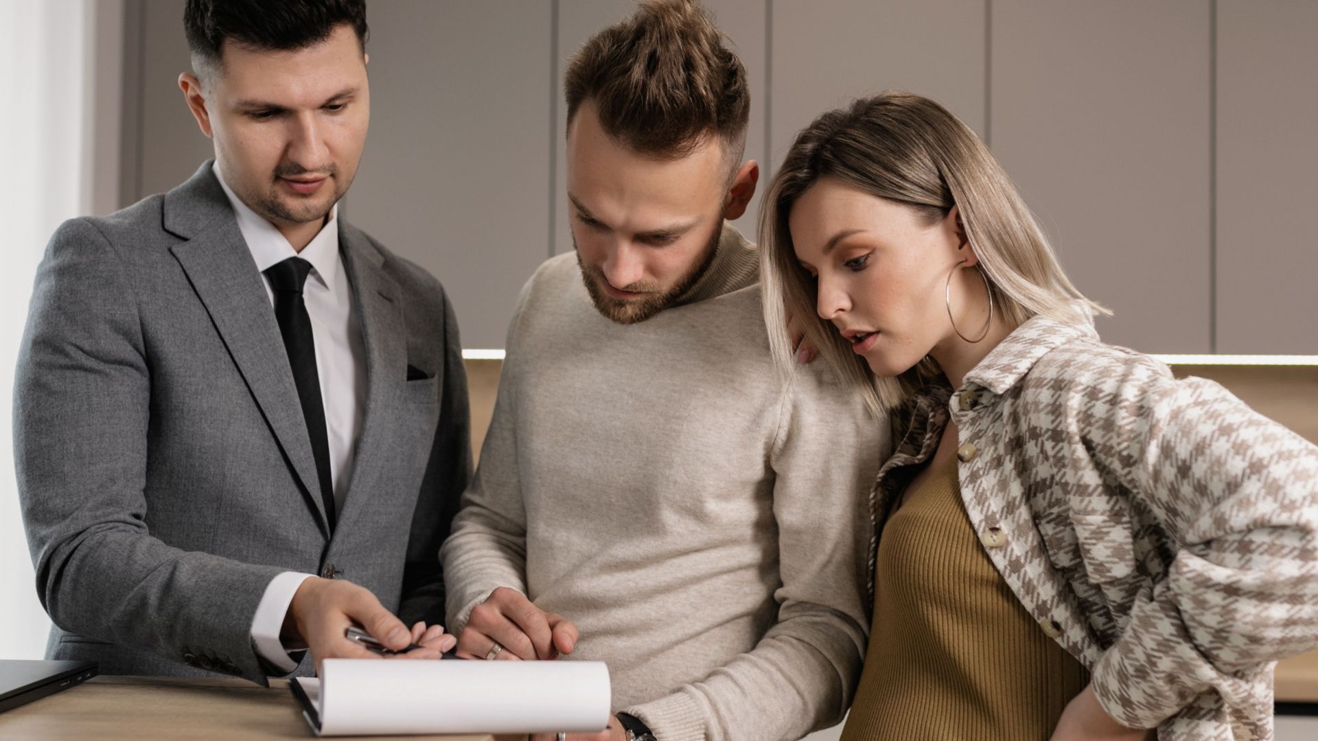 A young couple reviews and signs real estate documents with an agent in a modern kitchen setting.