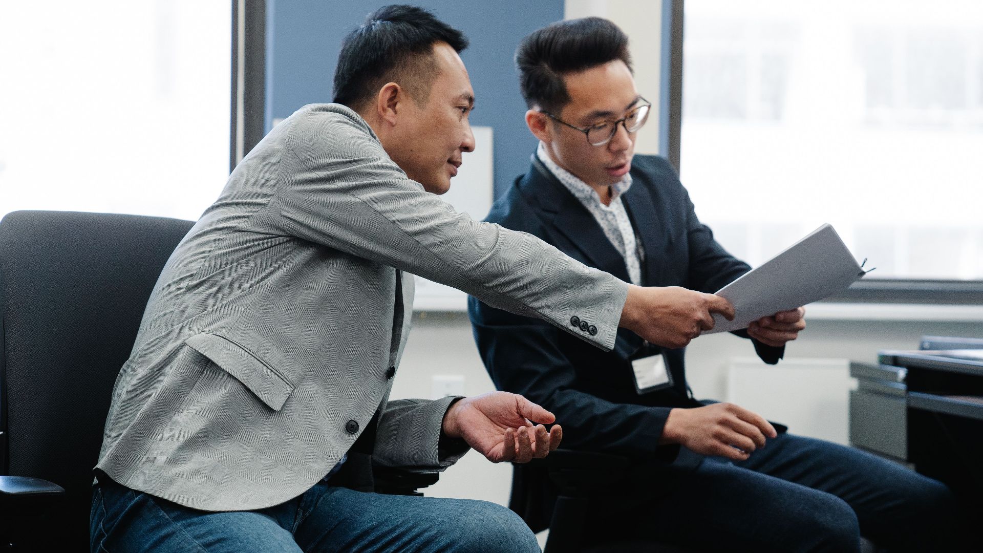 Two male colleagues exchanging documents in a modern office setting.